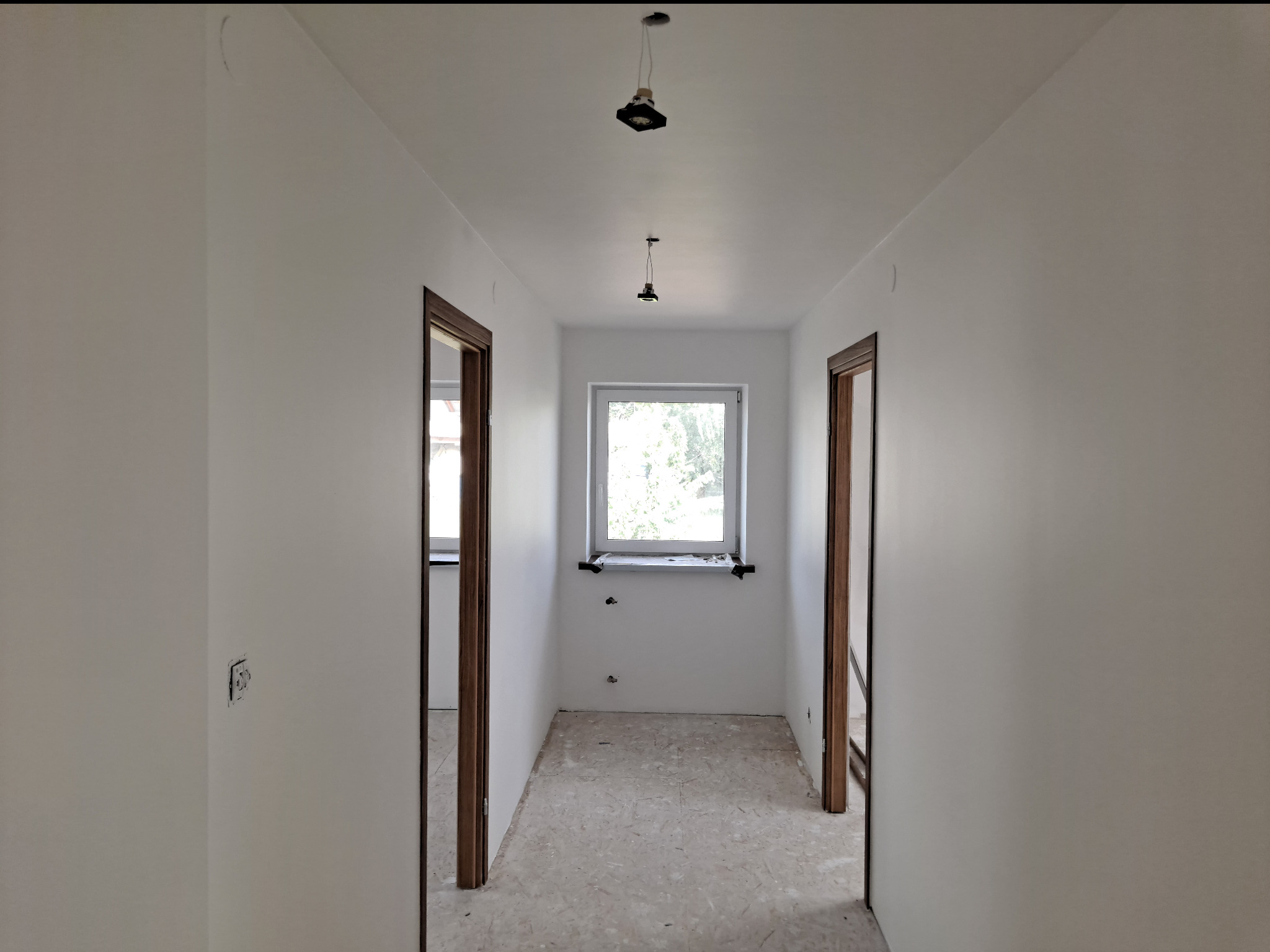 Korytarz w trakcie wykańczania, widoczne gładkie, białe ściany, osadzone ościeżnice drzwiowe bez skrzydeł, surowe podłoże podłogowe oraz wiszące przewody oświetleniowe.
