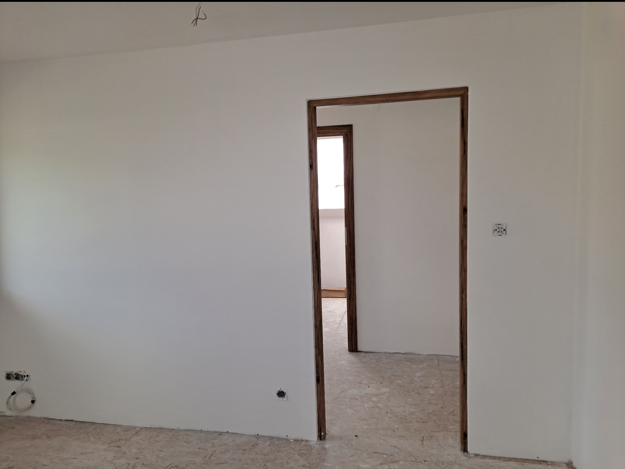 Wnętrze w trakcie prac wykończeniowych: białe ściany, widoczne gniazdka elektryczne, dwa drewniane ościeża drzwiowe prowadzące do kolejnych pomieszczeń, surowe podłoże podłogowe.