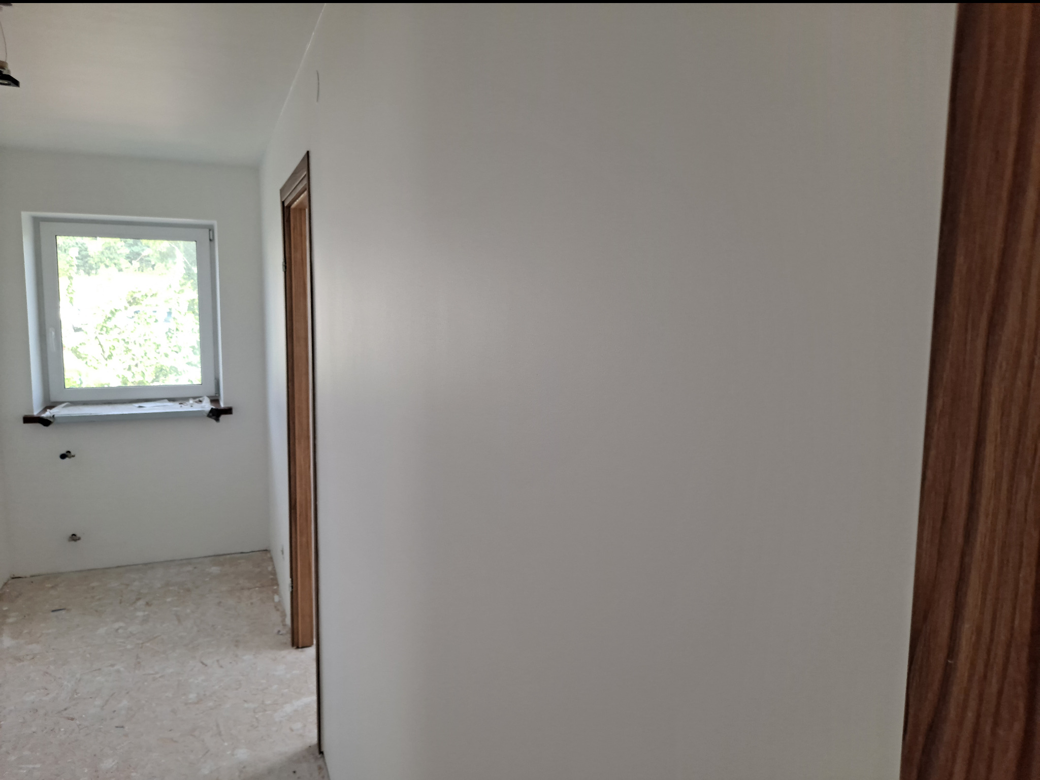 Wnętrze pokoju po świeżo wykonanych pracach malarskich, widoczna gładka, biała ściana, drewniana ościeżnica drzwi i okno z widokiem na zieleń.