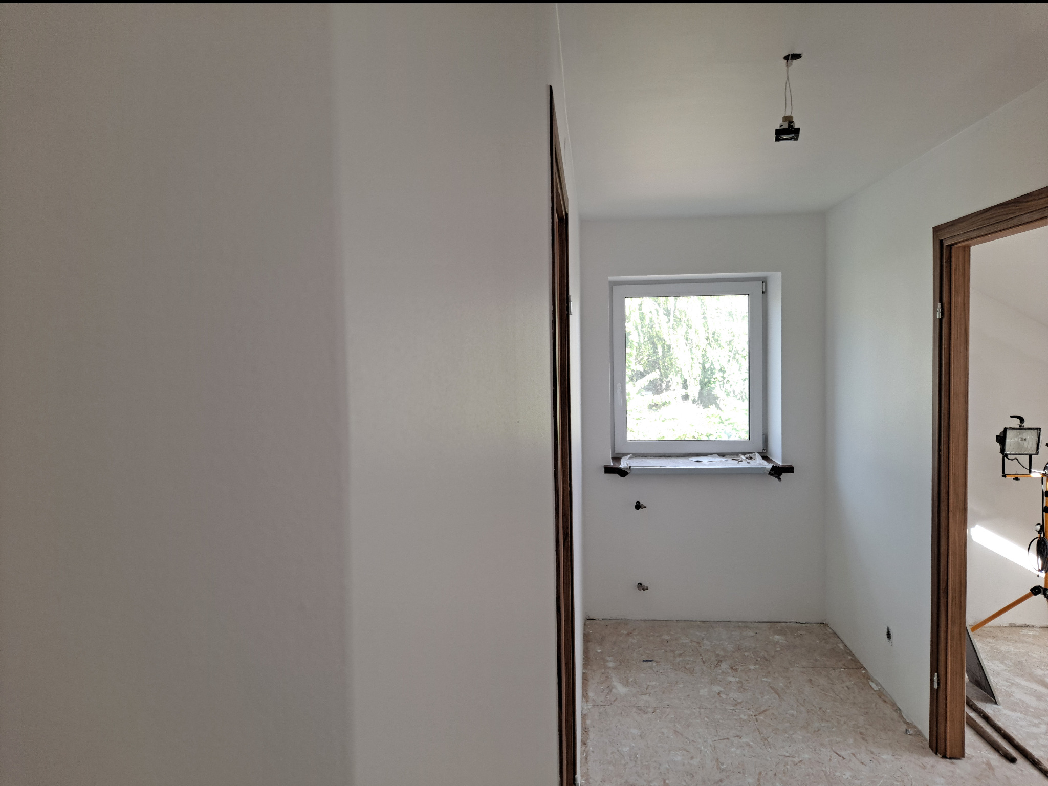 Wnętrze pokoju po malowaniu na biało, widoczne przygotowane ościeżnice drzwiowe, okno z parapetem i prowizoryczne oświetlenie na statywie.