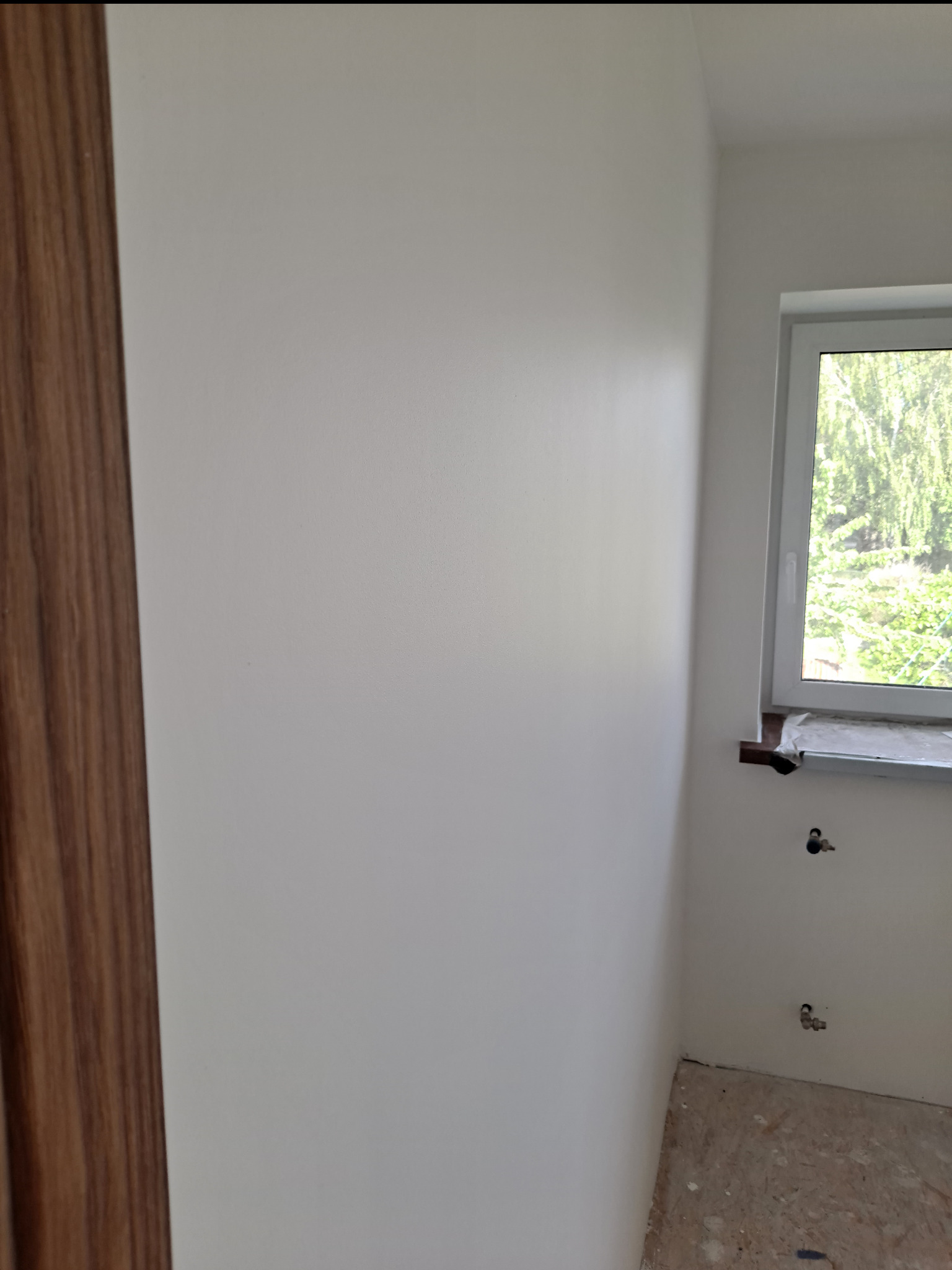 Świeżo pomalowane, jasne ściany w narożniku pokoju z oknem, widoczne ślady po pracach wykończeniowych na podłodze i wystające zawory grzejnikowe.