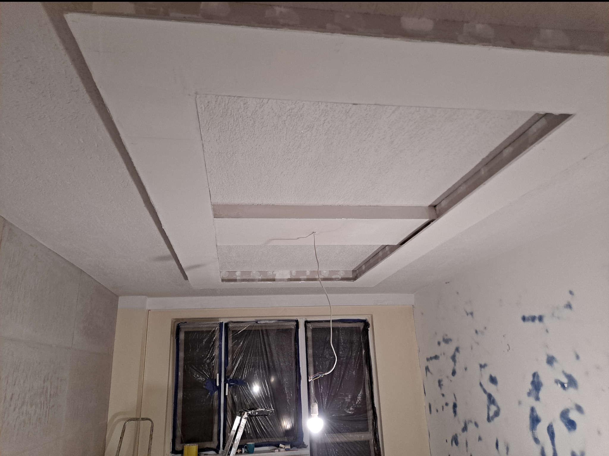 Biały sufit podwieszany z kwadratową wnęką i oświetleniem, widoczne przygotowania do prac wykończeniowych na ścianach i zabezpieczone okna folią.
