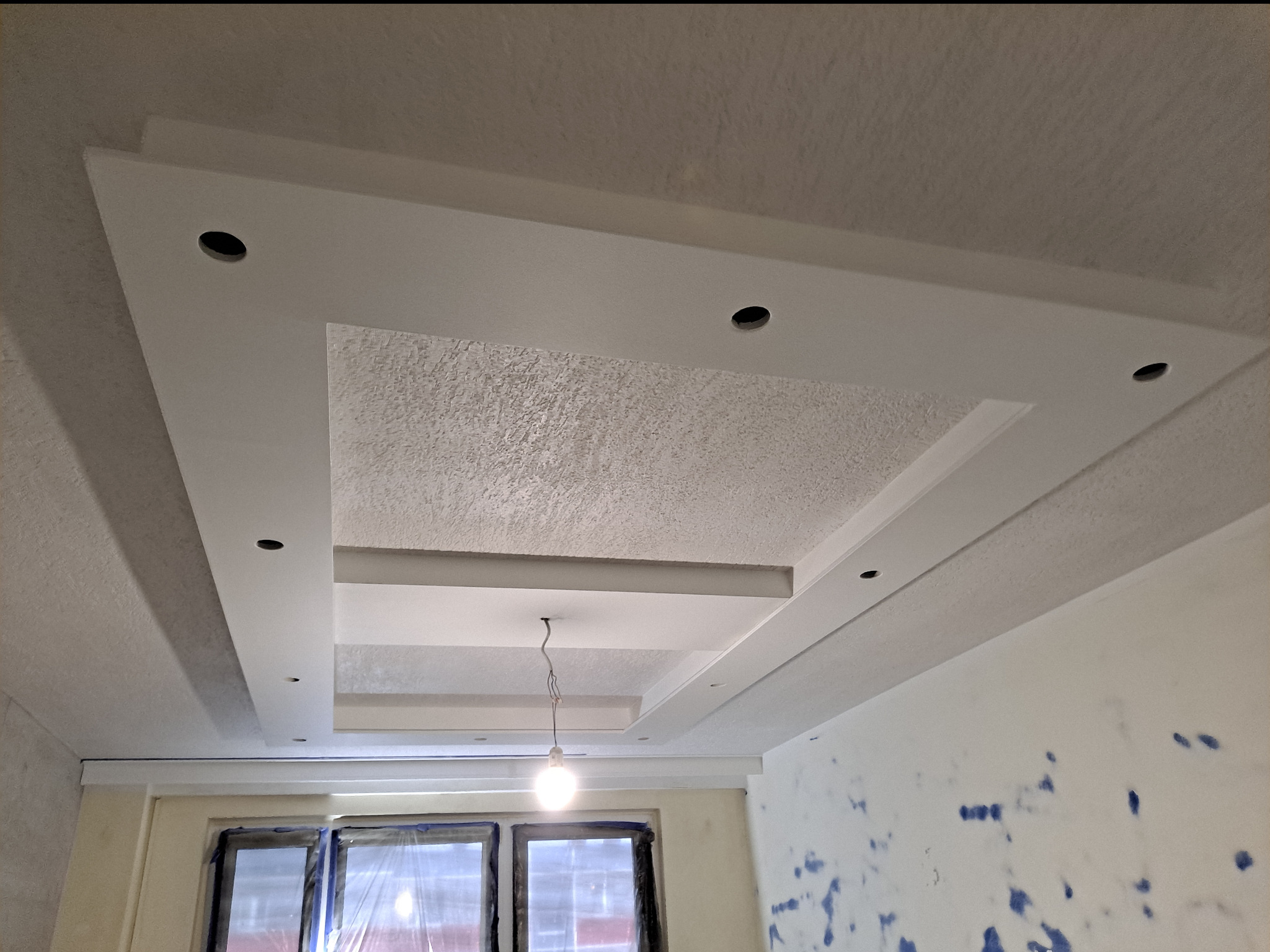 Kaskadowy sufit z gładzi z wnękami na oświetlenie punktowe i wiszącą żarówką na kablu, widoczne ślady po malowaniu ścian.