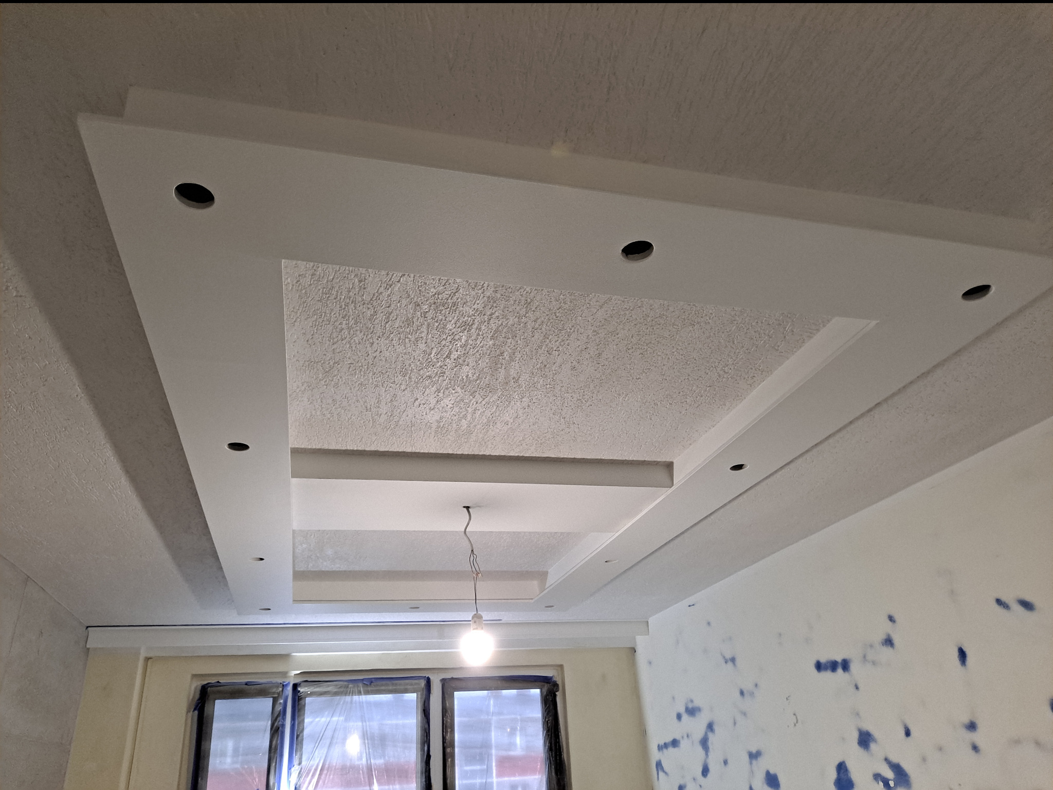 Wielopoziomowy, biały sufit podwieszany z oświetleniem punktowym w trakcie montażu, widoczne przewody zwisające z sufitu, ściany z plamami farby, okna zabezpieczone folią.