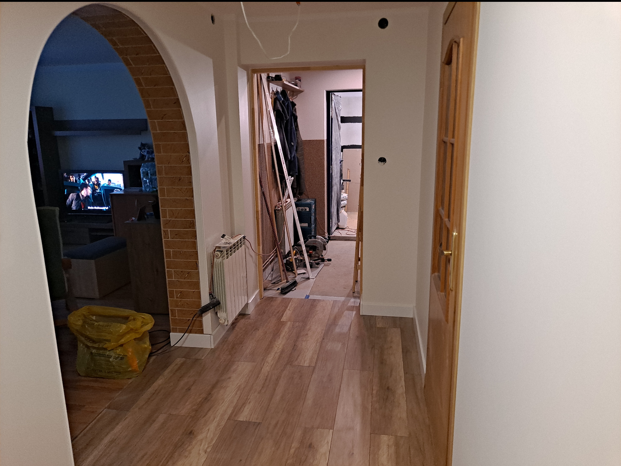 Wnętrze domu w trakcie prac wykończeniowych: świeżo ułożona podłoga z paneli, widoczne narzędzia i materiały budowlane w przejściu, żółty worek z odpadami, grzejnik, oraz widoczny fragment...