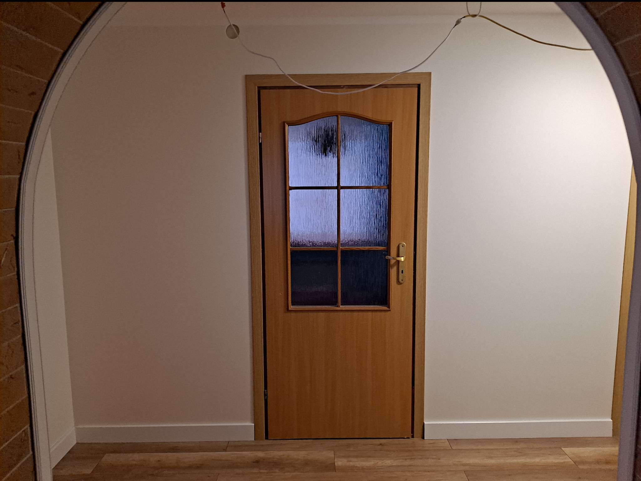 Drewniane drzwi wewnętrzne z szybami o fakturze szkła ornamentowego, umieszczone w białej ścianie, widoczne z perspektywy łuku wejściowego z ceglaną okładziną.