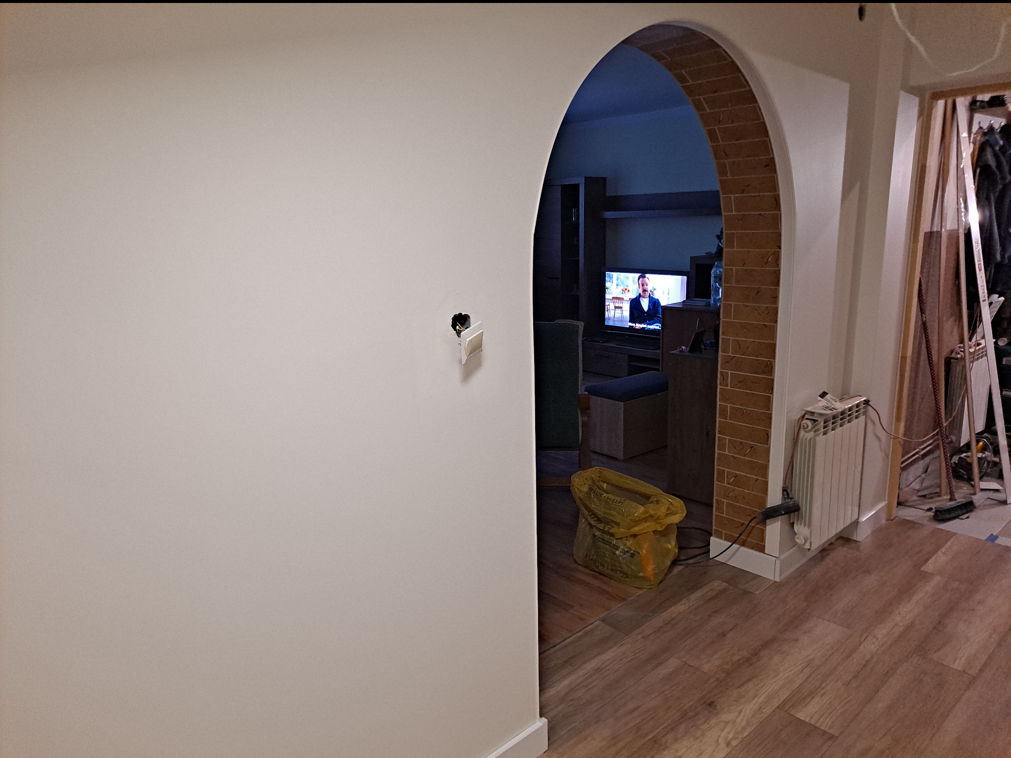 Wnętrze mieszkania w trakcie prac wykończeniowych: gładka, biała ściana z niedokończonym gniazdkiem elektrycznym, łukowe przejście do pokoju z telewizorem, grzejnik i worek na śmieci na drewnianej...