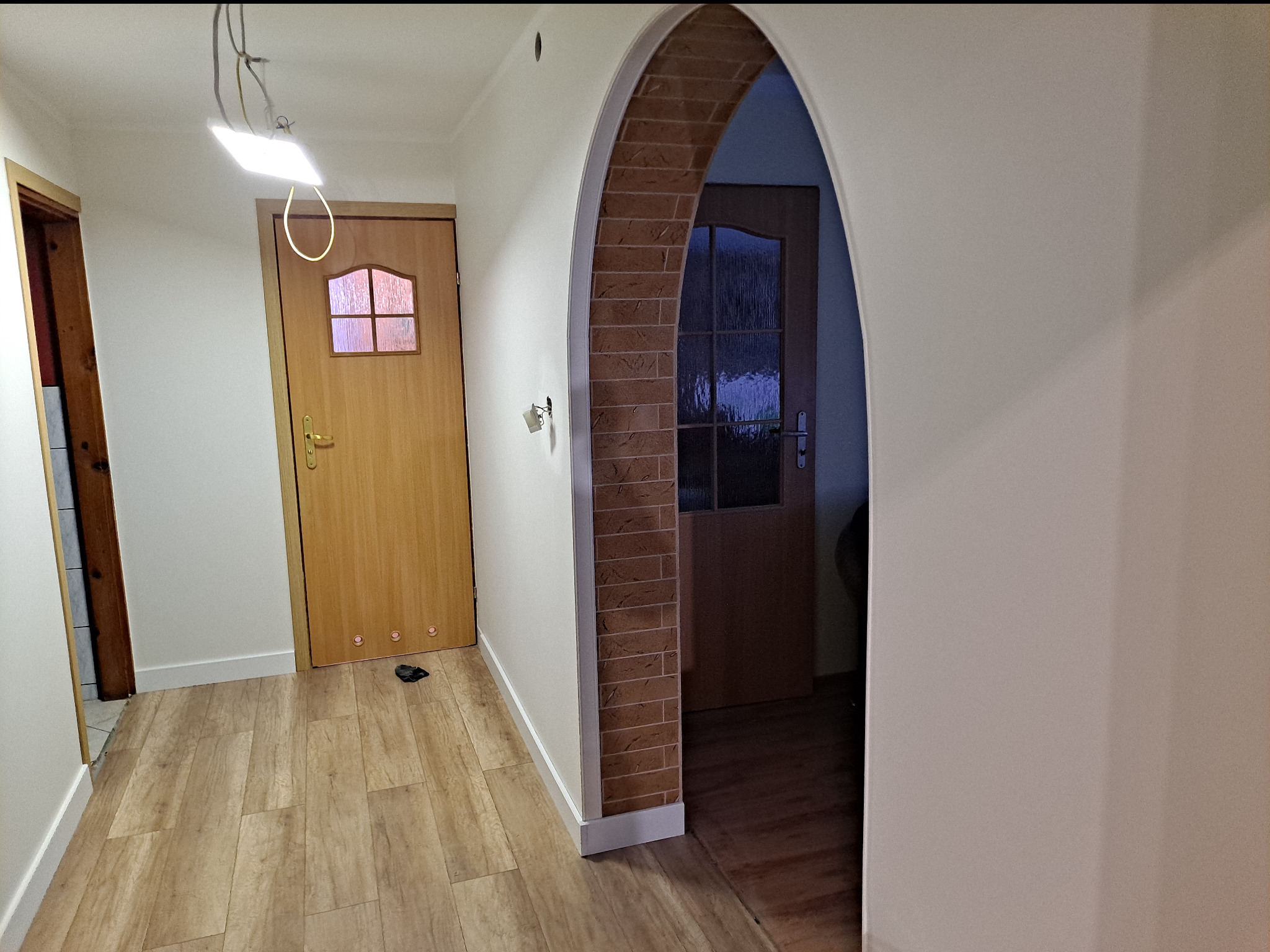 Korytarz z drewnopodobną podłogą, dwoje drzwi z ościeżnicami, jedno z ceglanym łukiem, surowa instalacja oświetleniowa na suficie.