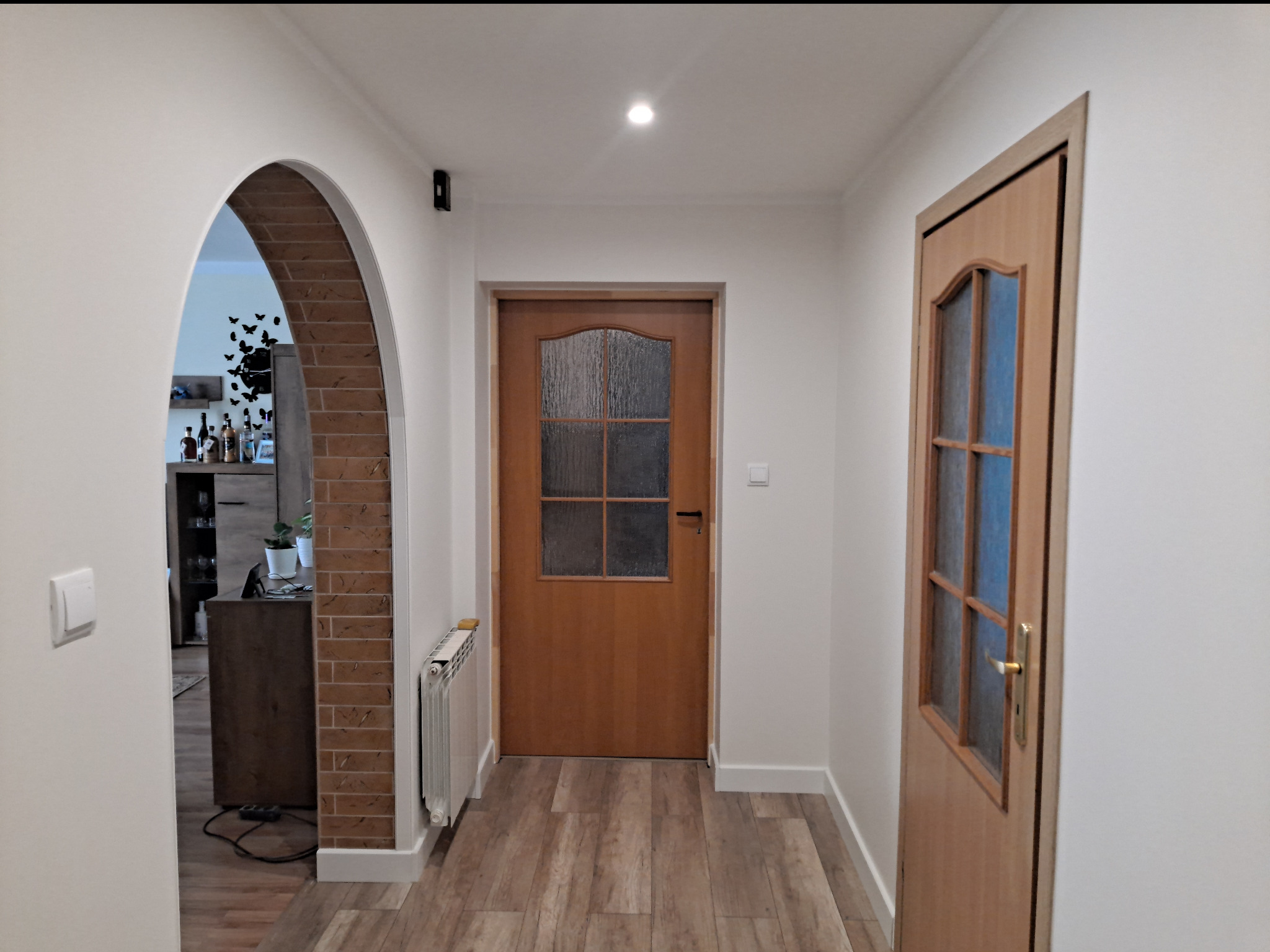 Korytarz mieszkania po remoncie z dwiema drewnianymi drzwiami z szybami, łukowate wejście do pokoju z ceglanym obramowaniem, grzejnik i panele podłogowe imitujące drewno.