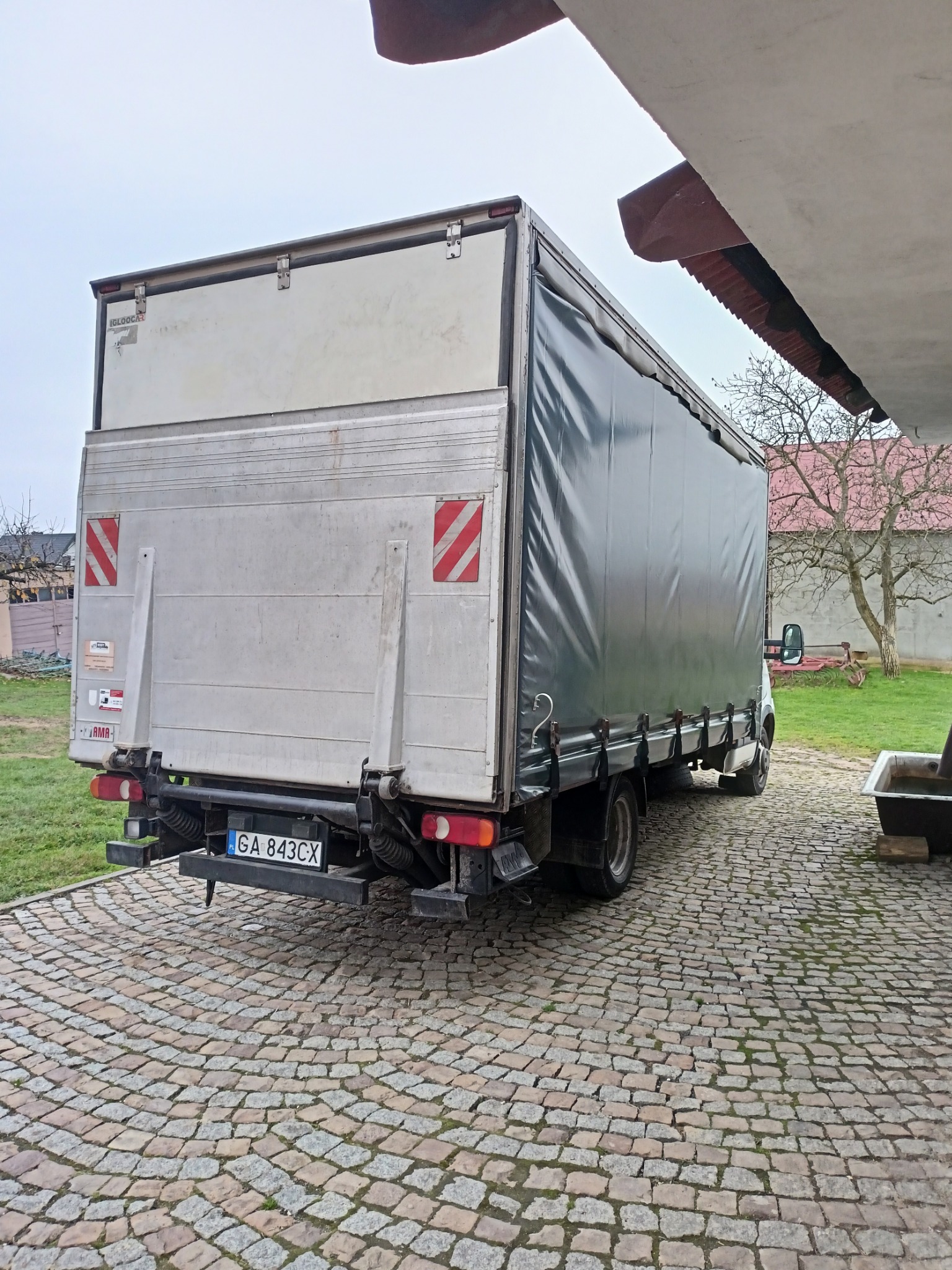 Tył samochodu dostawczego Iveco z plandeką, zaparkowanego na brukowanej nawierzchni. Widoczne tablice rejestracyjne GA 843CX, czerwono-białe pasy ostrzegawcze na tylnych drzwiach i fragment windy...