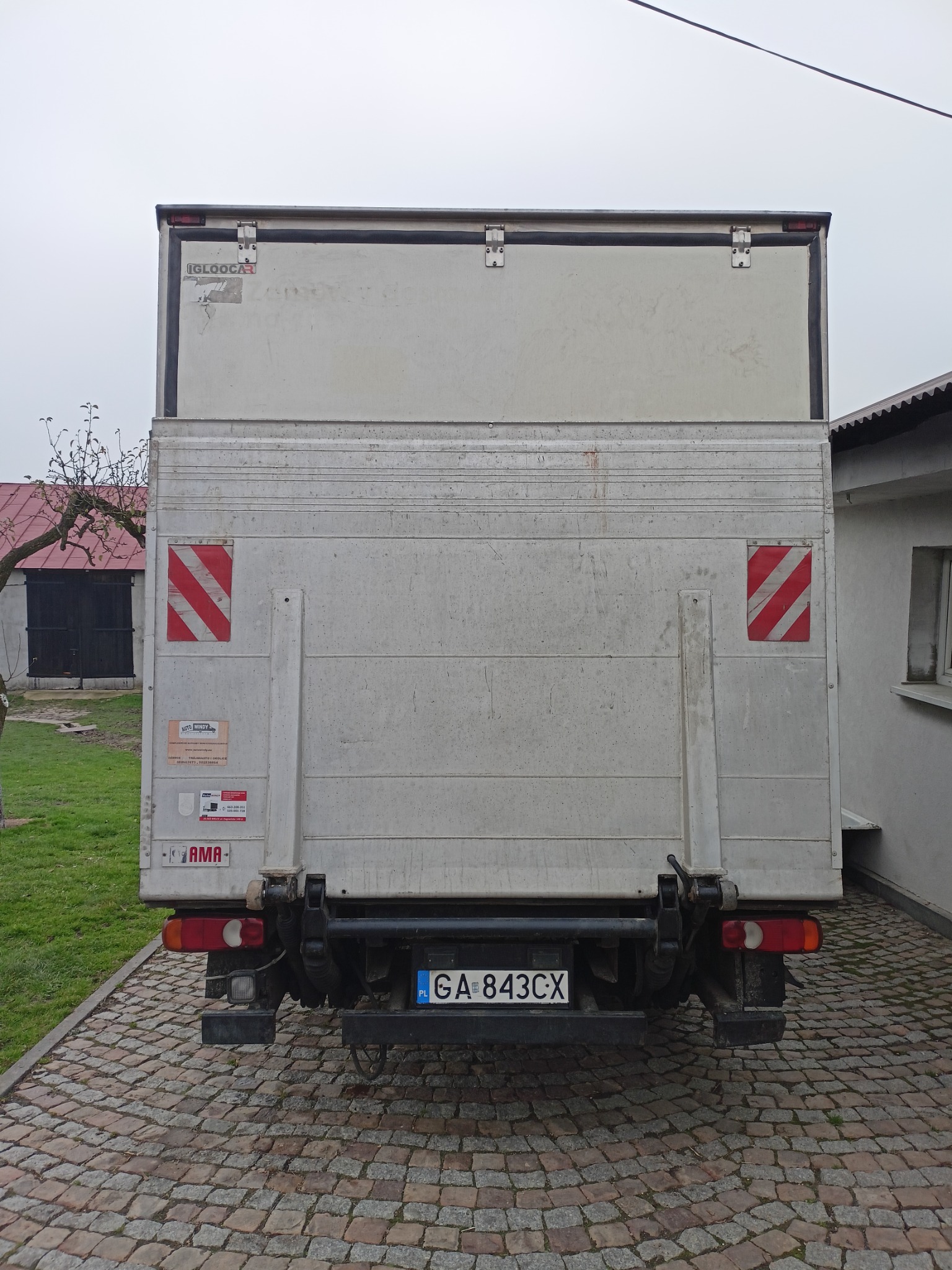 Tył samochodu dostawczego z zabudową kontenerową, widoczne tablice ostrzegawcze, tablica rejestracyjna i elementy oświetlenia, zaparkowany na brukowanym podjeździe.