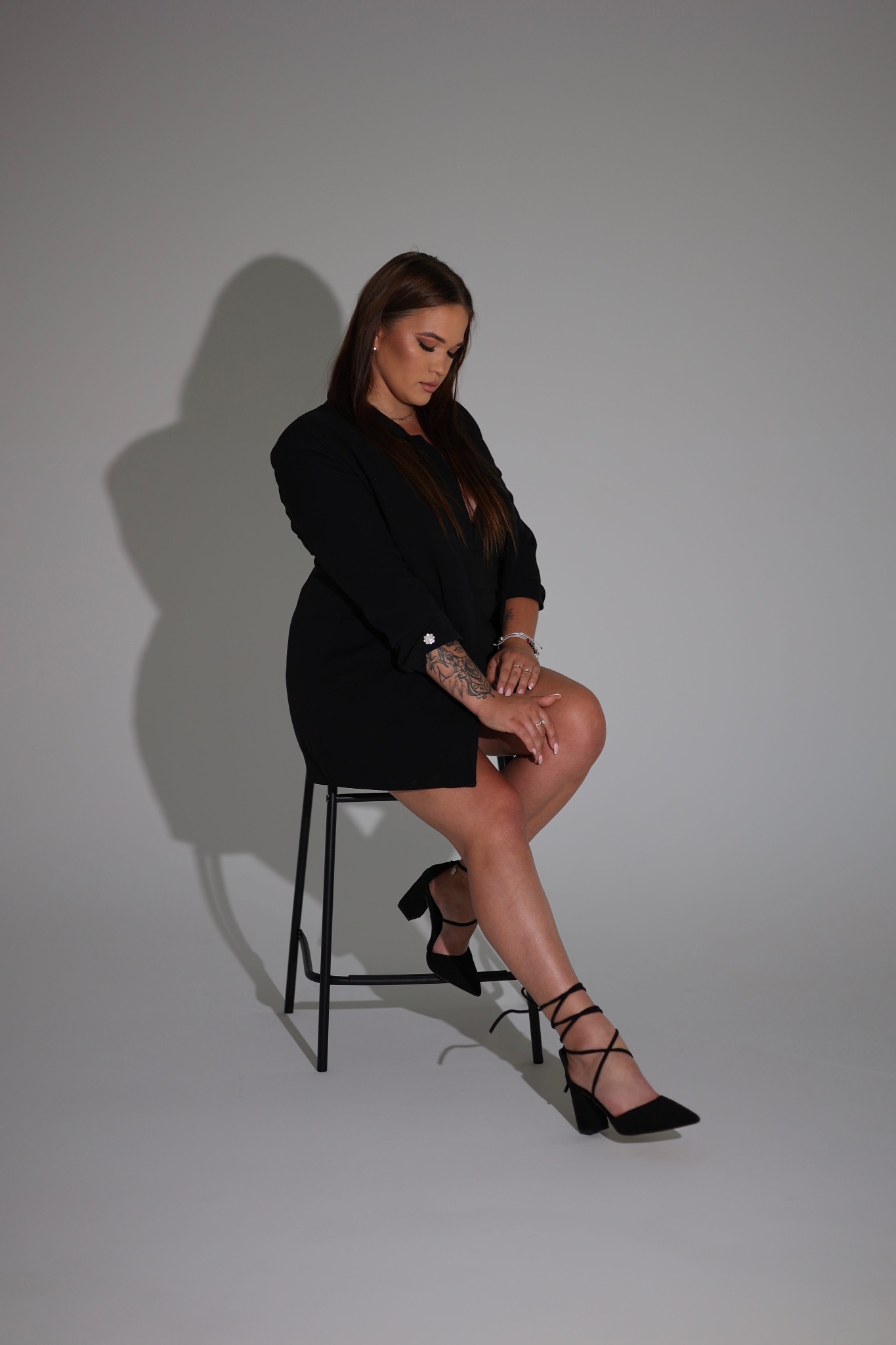 Kobieta w czarnej sukience siedzi na stołku na szarym tle. Widoczne tatuaże na ramieniu i eleganckie czarne buty na obcasie ze sznurowaniem.
