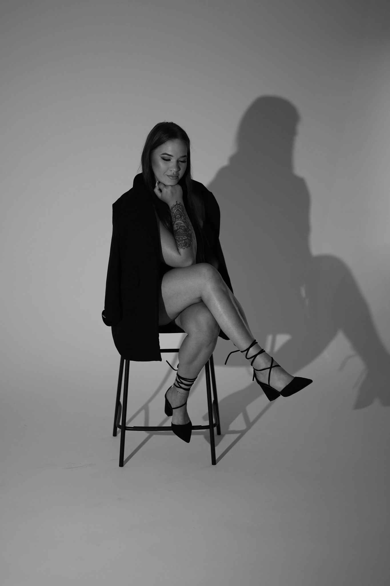 Czarno-biała fotografia kobiety z tatuażem, siedzącej na krześle w studiu w Poznaniu. Ubrana w marynarkę, spogląda w dół. Oświetlenie tworzy wyraźny cień.