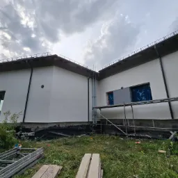 Biała elewacja domu w trakcie prac wykończeniowych, widoczne rusztowania i narzędzia. Częściowo zakryte okna folią ochronną. Pochmurne niebo w tle.