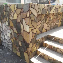 Kamienna okładzina elewacyjna na schodach wejściowych, nieregularne płytki w odcieniach beżu, brązu i szarości, fuga w kolorze zbliżonym do kamienia, widoczne materiały budowlane na górze konstrukcji.