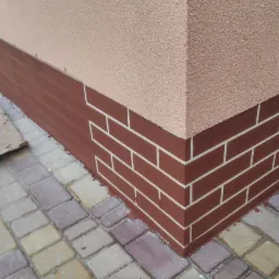 Narożnik budynku z elewacją pokrytą beżowym tynkiem strukturalnym i cokołem z brązowej imitacji cegły z białymi fugami, widoczne kostki brukowe na chodniku.
