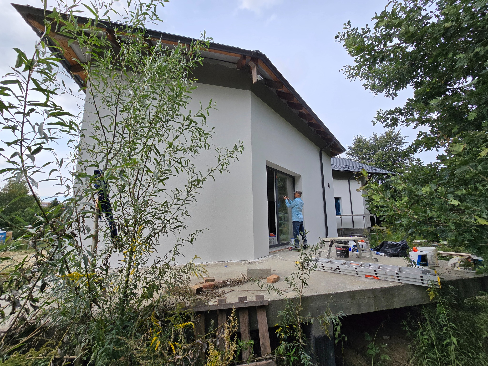 Elewacja domu jednorodzinnego w trakcie wykończenia, widoczny tynkarz przy oknie, drabina i narzędzia. Dom o nowoczesnej architekturze, otoczony zielenią.