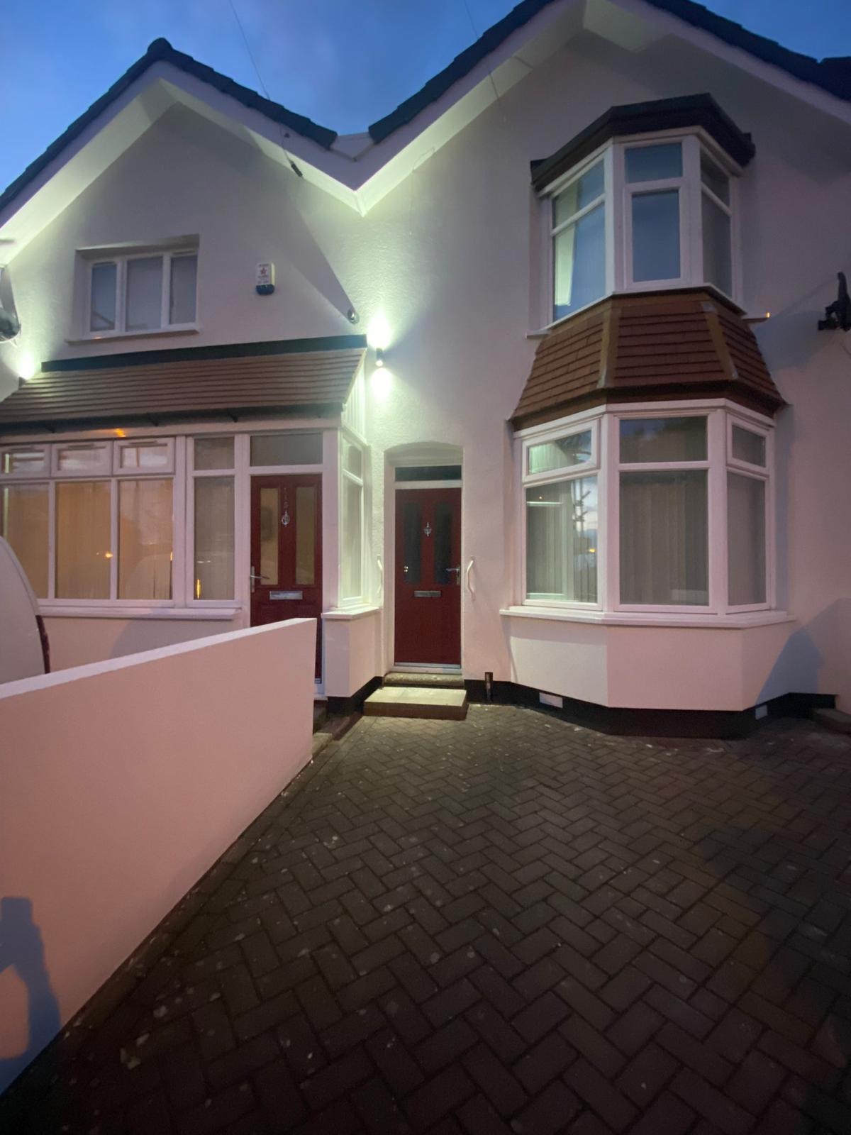 Biały dom z czerwonymi drzwiami wejściowymi i oświetleniem elewacji, widok z brukowanego podjazdu o zmierzchu. Charakterystyczne okna wykuszowe.