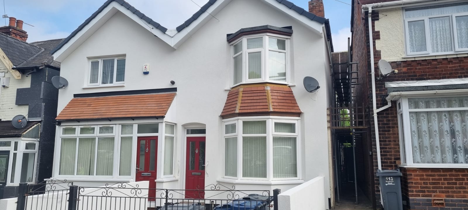 Biały dom z czerwonymi drzwiami i okiennicami, widok z przodu. Widoczne rusztowania z boku budynku, sugerujące prace elewacyjne.