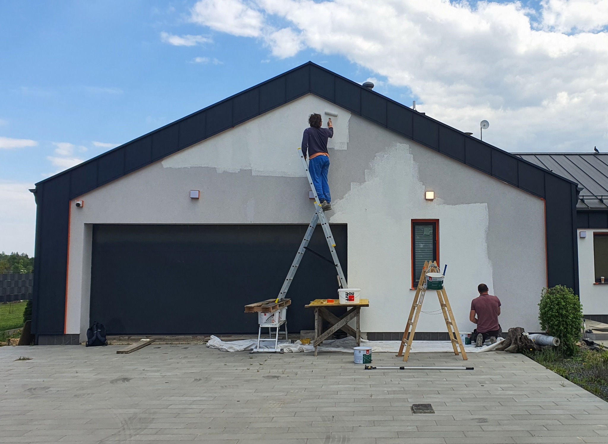 Dwóch mężczyzn maluje elewację domu jednorodzinnego, jeden na drabinie z wałkiem, drugi klęczy przy ścianie z pędzlem, widoczne narzędzia malarskie i wiaderka z farbą.