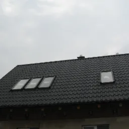 Czarny dach z dachówki modułowej z trzema oknami dachowymi po lewej stronie i jednym oknem dachowym po prawej stronie, widok z dołu na tle pochmurnego nieba.