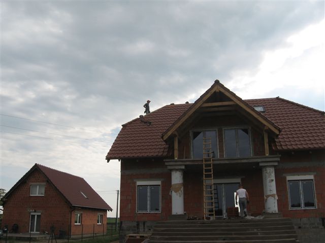 Budowa domu jednorodzinnego z czerwonej cegły z dachem pokrytym czerwoną dachówką; na dachu pracuje dekarz, a na balkonie stoi drabina.