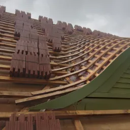 Konstrukcja dachu z drewnianych łat i kontrłat, częściowo pokryta brązową dachówką karpiówką, widoczne pakiety dachówek przygotowane do ułożenia, pochmurne niebo w tle.