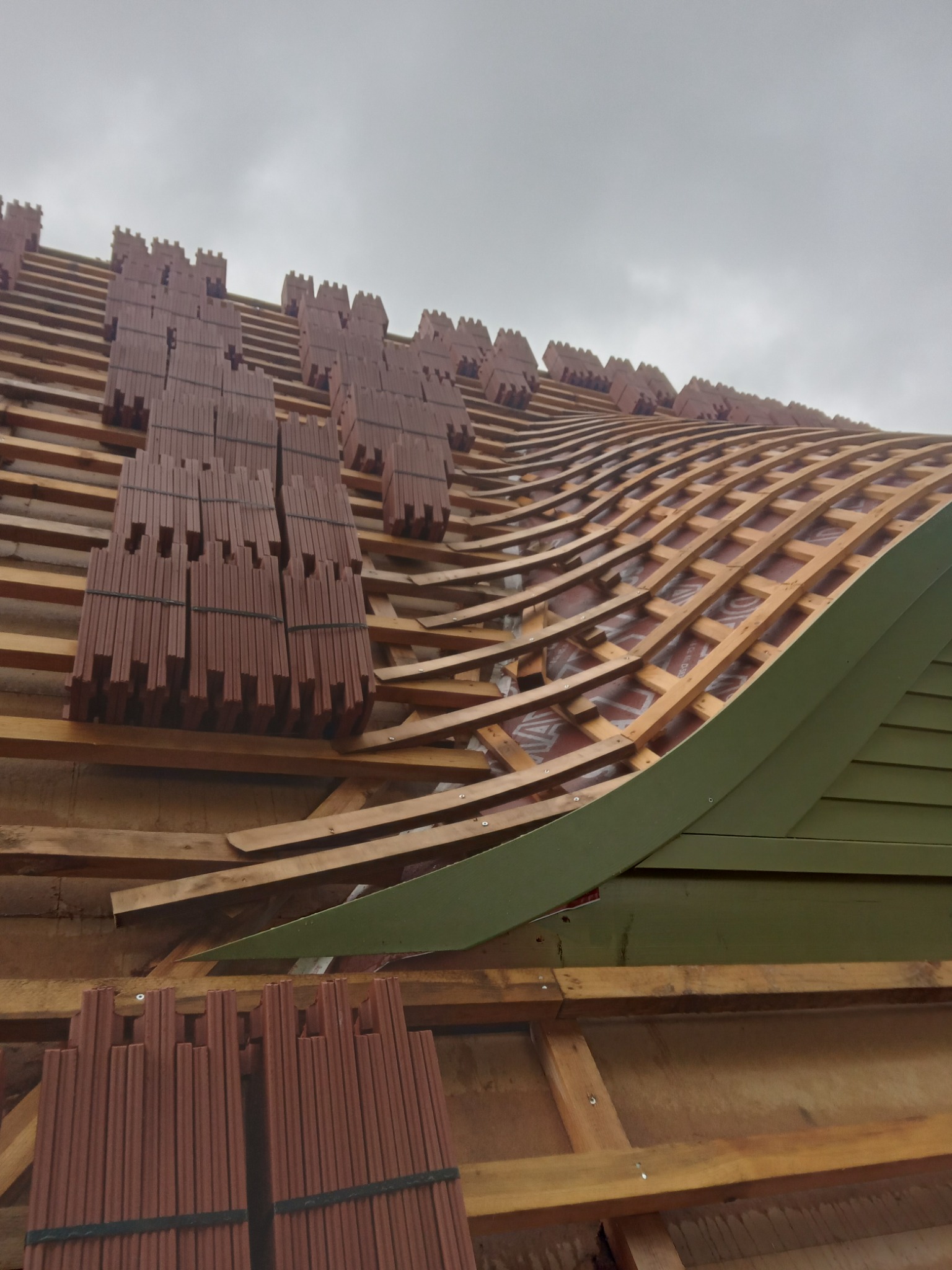 Konstrukcja dachu z drewnianych łat i kontrłat, częściowo pokryta brązową dachówką karpiówką, widoczne pakiety dachówek przygotowane do ułożenia, pochmurne niebo w tle.