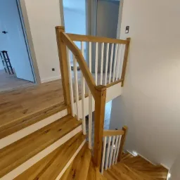 Spiralne schody z jasnego drewna z białymi podstopnicami i balustradą, widok z góry, w tle drzwi z szarą ościeżnicą.