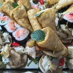 Różnorodny wybór przekąsek na tacy: sushi z łososiem, roladki z ciasta z ziołami i pastą, kanapeczki z pastą i grzybami, udekorowane listkami mięty i papryczką.