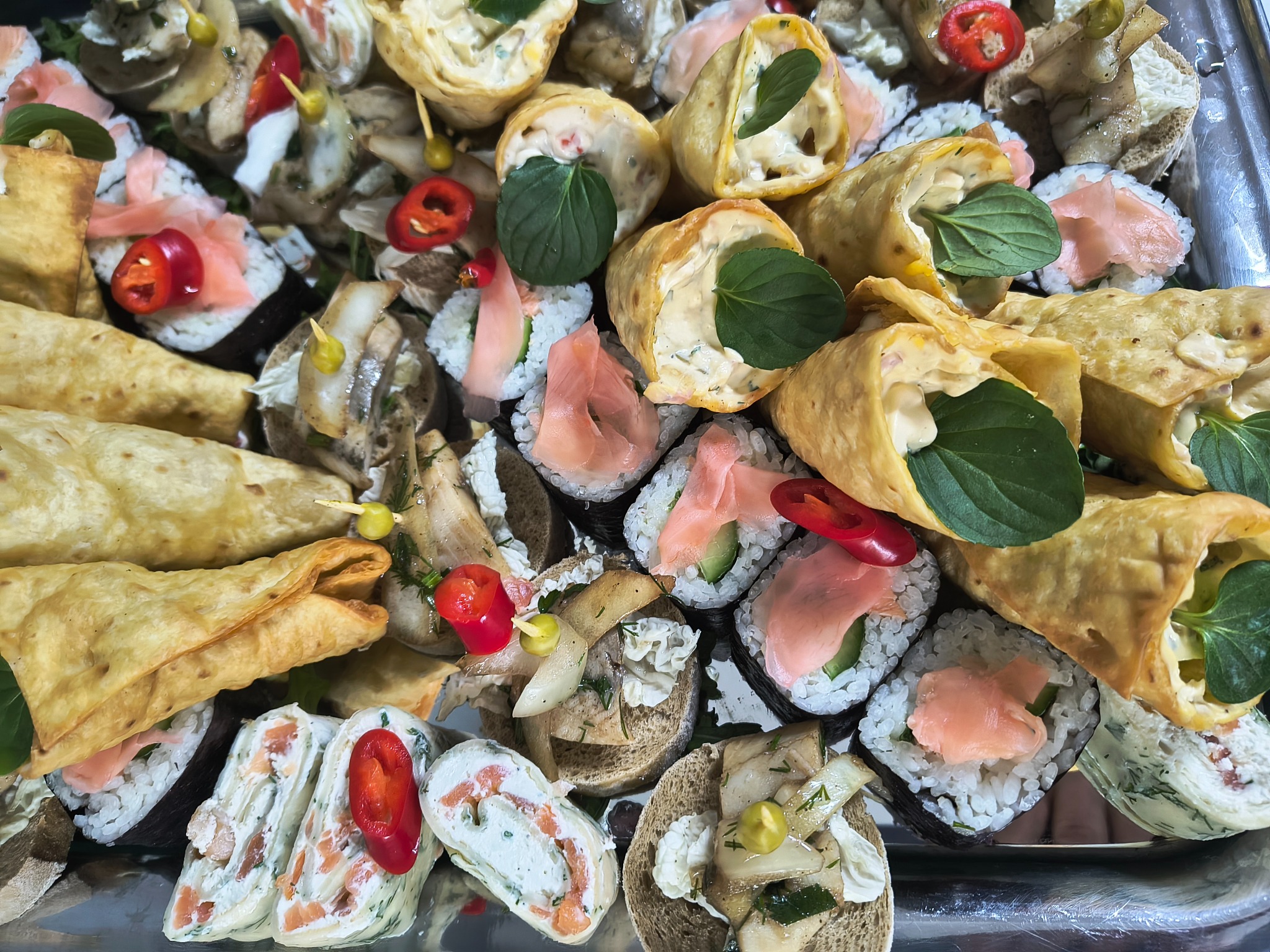 Zestaw różnorodnych przekąsek na srebrnej tacy: sushi z łososiem i imbirem, roladki omletowe z nadzieniem, mini kanapeczki z marynowanym śledziem i papryczką chili, udekorowane listkami mięty.
