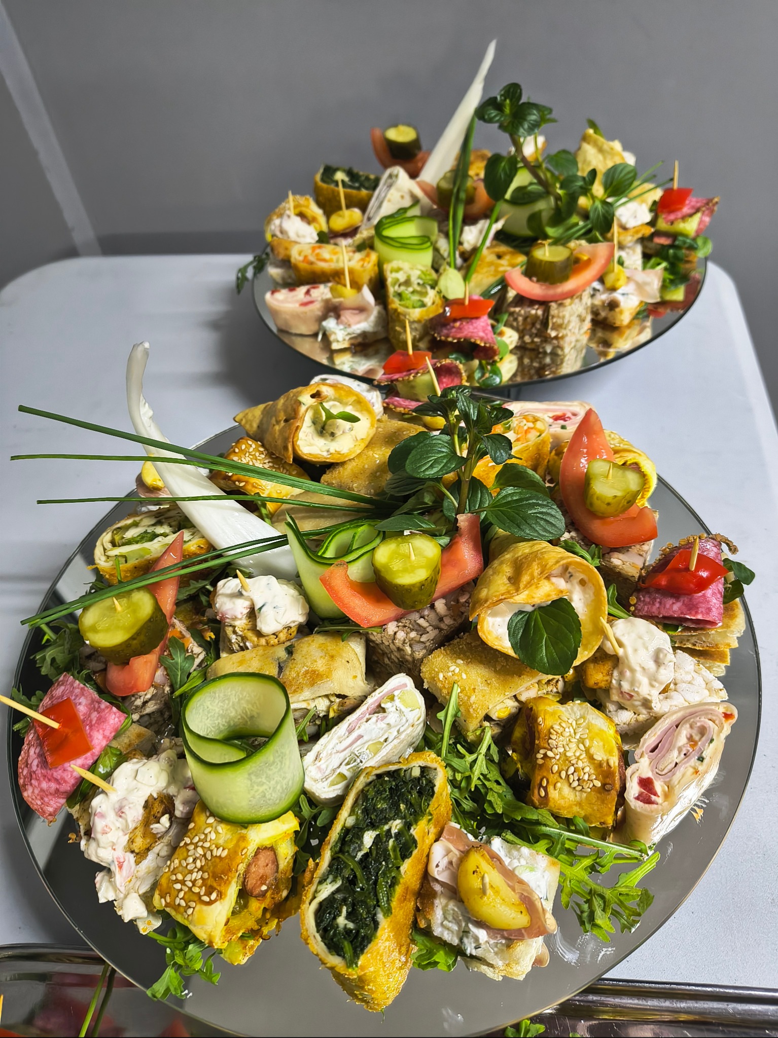 Dwa okrągłe, lustrzane talerze z różnorodnymi, kolorowymi przekąskami na przyjęcie, udekorowane świeżymi ziołami i warzywami, z widocznymi roladkami, koreczkami i tartinkami.