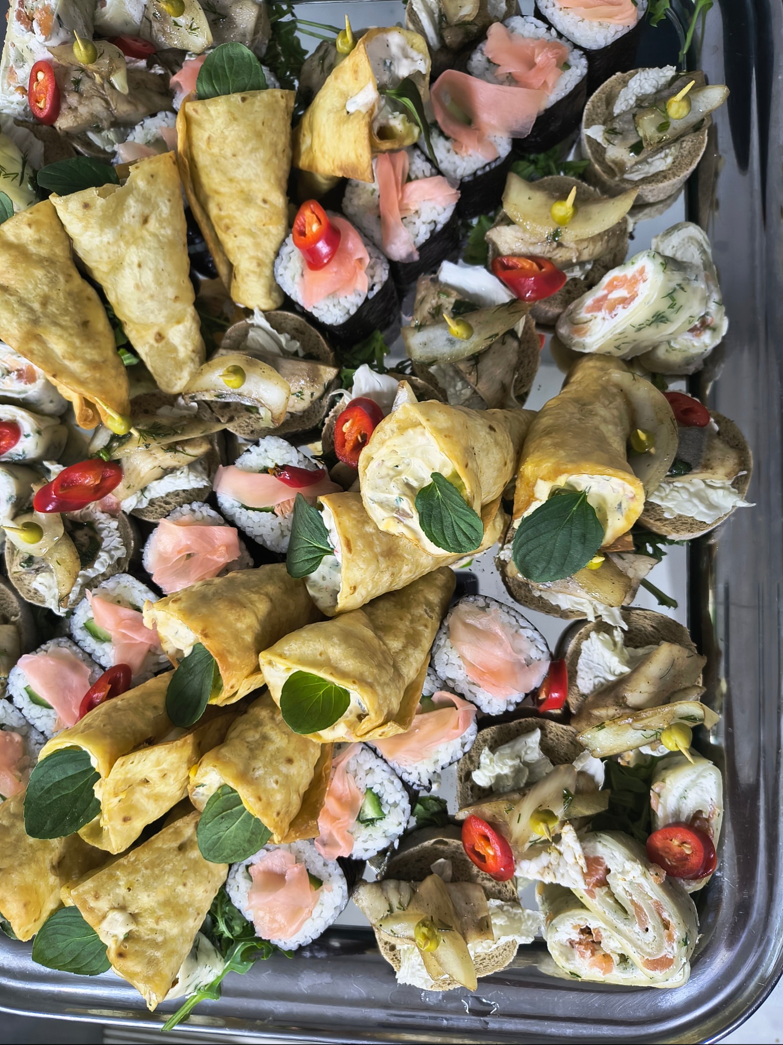 Srebrna taca z różnorodnymi przekąskami: roladki z tortilli z zielonymi listkami, sushi z łososiem i kanapki z pastą, udekorowane kawałkami czerwonej papryczki chili.