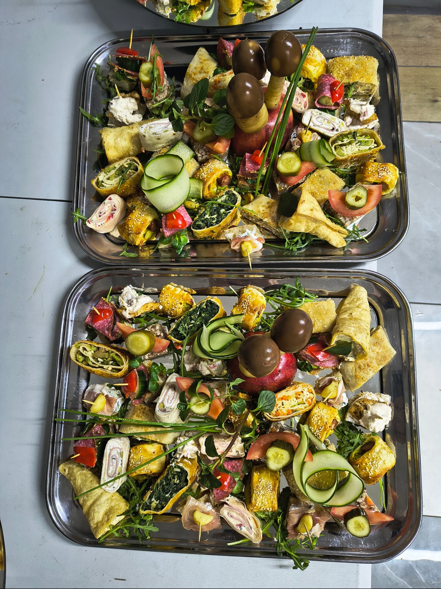 Dwie srebrne tace z różnorodnymi przekąskami: roladki z nadzieniem szpinakowym, tortille, koreczki z warzywami i wędlinami, dekorowane ogórkiem i świeżymi ziołami, z czekoladowymi grzybkami...