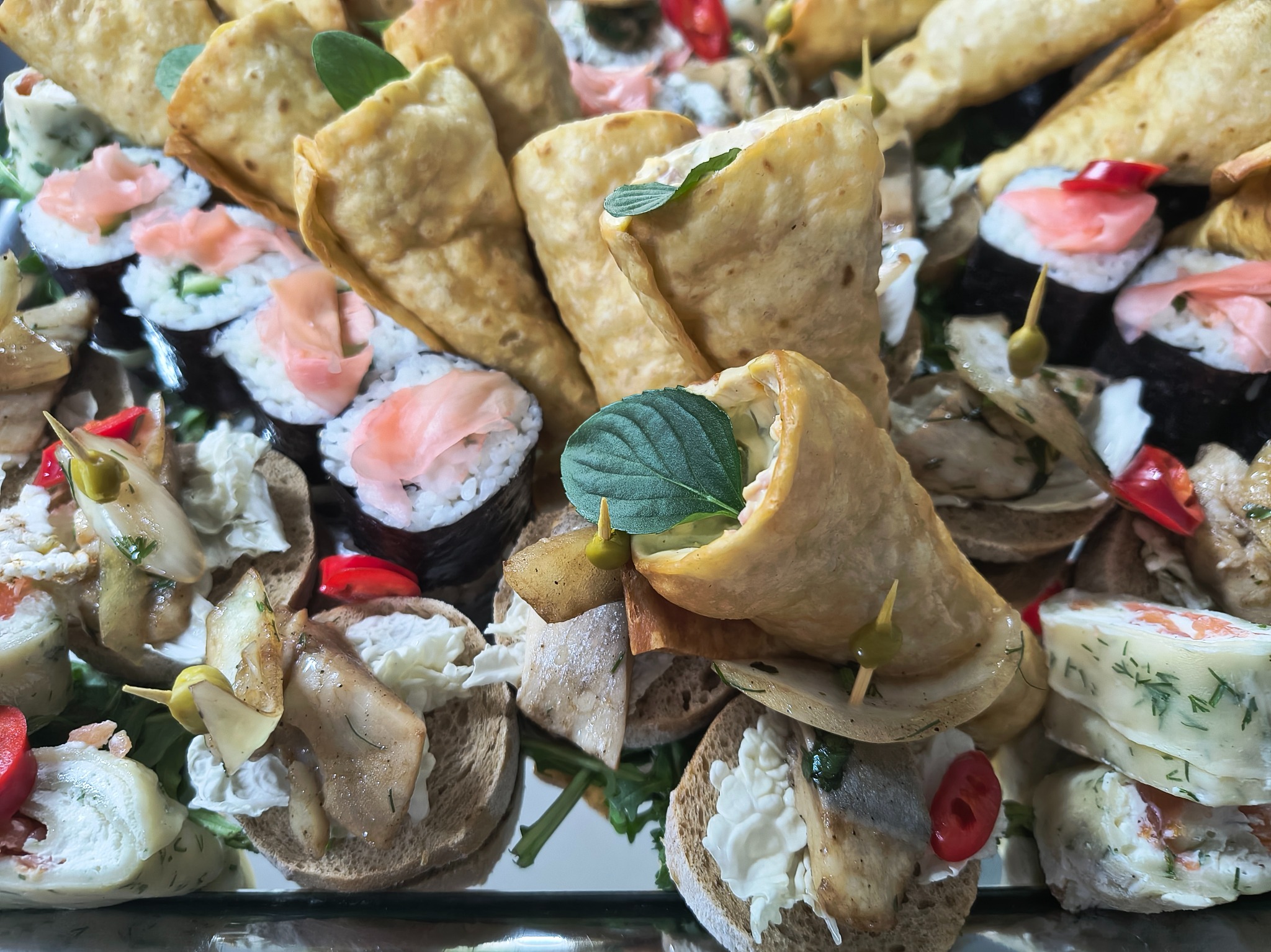 Różnorodny wybór przekąsek na tacy: sushi z łososiem, roladki z ciasta z ziołami i pastą, kanapeczki z pastą i grzybami, udekorowane listkami mięty i papryczką.