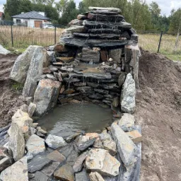 Kaskada wodna z kamienia łupkowego w Smardzewie, otoczona ziemią i większymi głazami, w tle dom jednorodzinny i ogrodzenie.