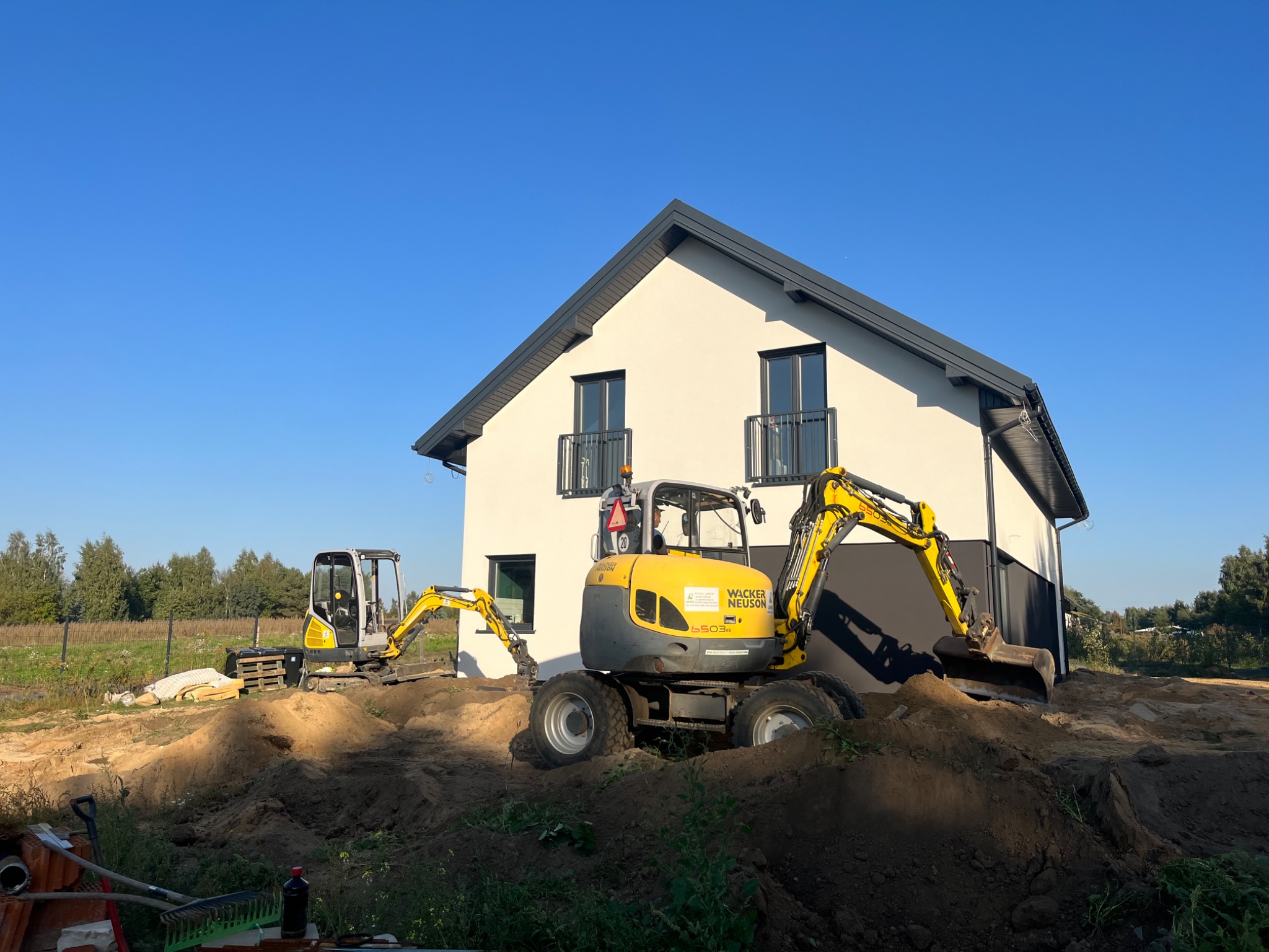Dwa żółte minikoparki Wacker Neuson na placu budowy przed nowoczesnym domem z białymi ścianami i ciemnoszarym dachem, ziemia nierówna, widoczne ślady pracy maszyn.