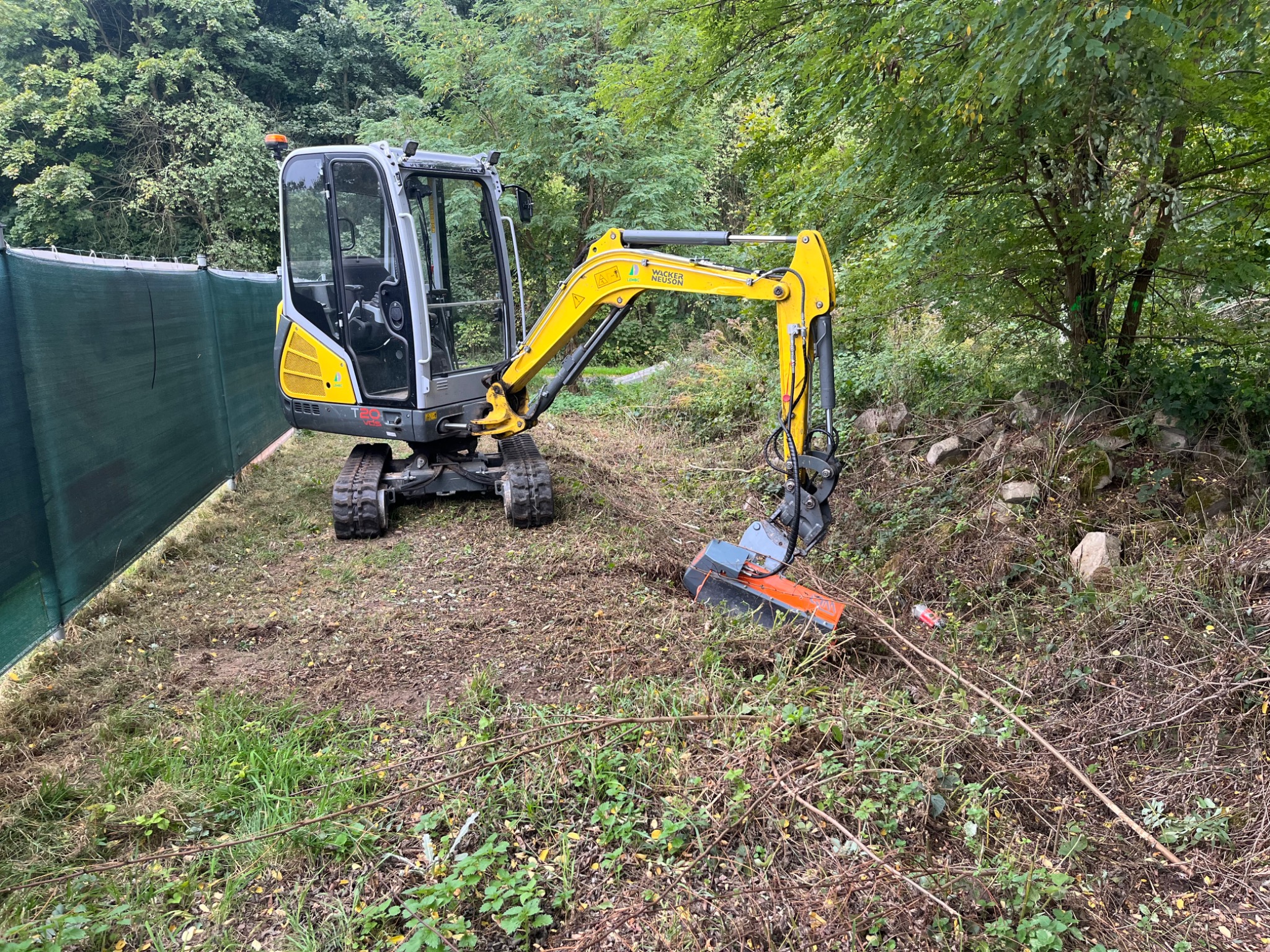 Żółto-szara mini koparka z frezem mulczującym na nierównym terenie z zieloną siatką w tle. Widoczne ścięte gałęzie i resztki roślinności.