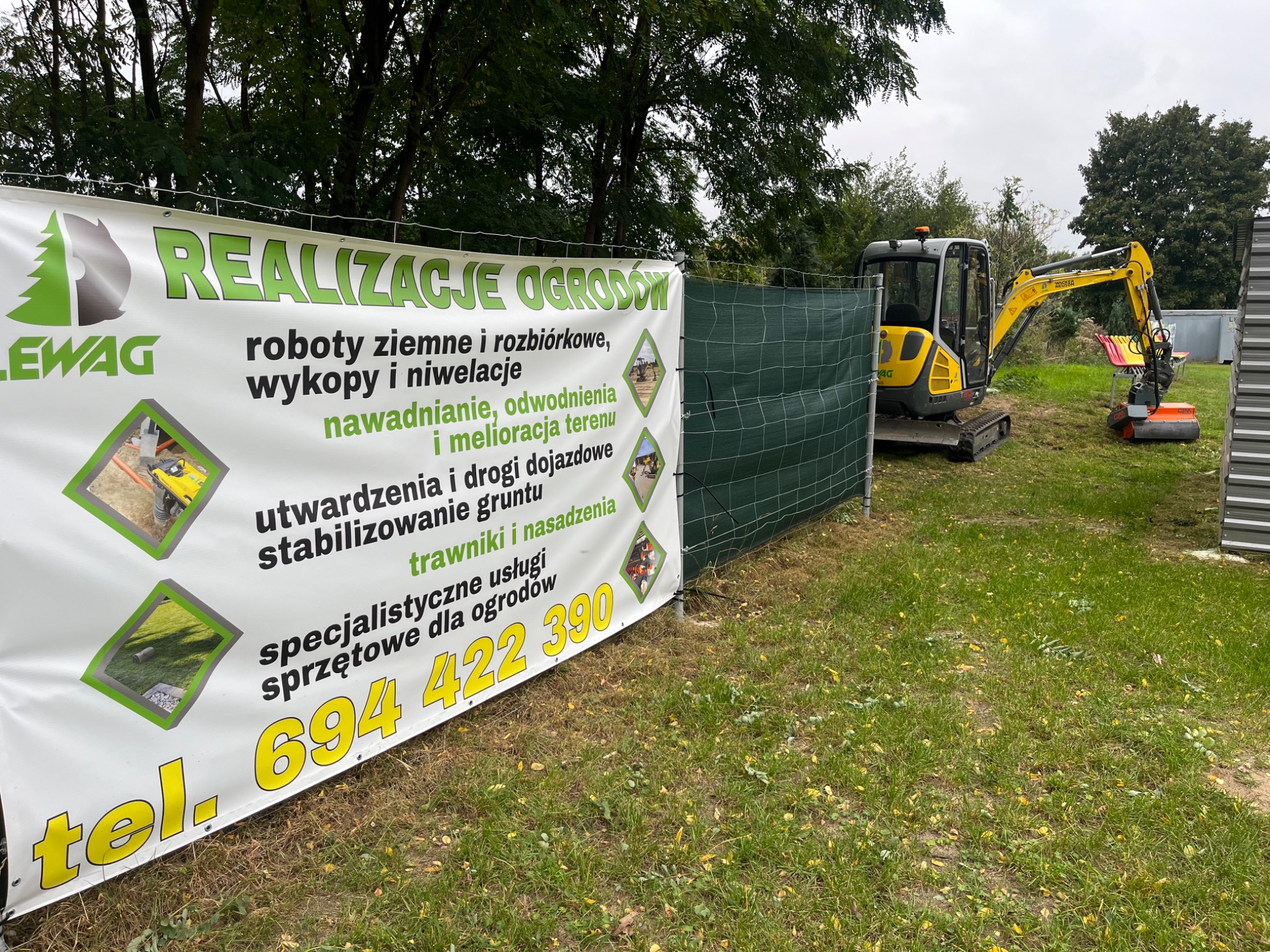Baner reklamowy firmy LEWAG z ofertą realizacji ogrodów, obejmującą roboty ziemne, wykopy, niwelacje, nawadnianie, utwardzanie gruntu i trawniki, na tle zielonej siatki ogrodzeniowej i żółtej mini...