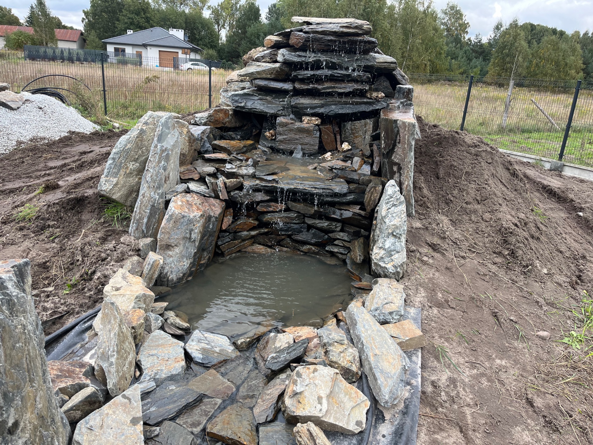 Kaskada wodna z kamienia łupkowego w Smardzewie, otoczona ziemią i większymi głazami, w tle dom jednorodzinny i ogrodzenie.