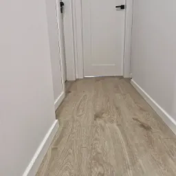 Jasne panele podłogowe imitujące drewno w korytarzu z białymi ścianami i białymi drzwiami z czarnymi klamkami.