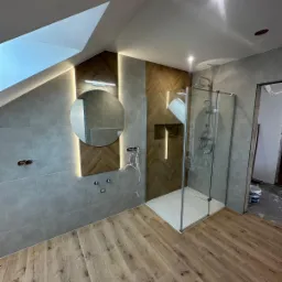 Wykończona łazienka na poddaszu: kabina prysznicowa, okrągłe lustro z podświetleniem LED, drewnopodobne płytki na podłodze i ścianie z wzorem jodełki. Częściowo widoczne kable.