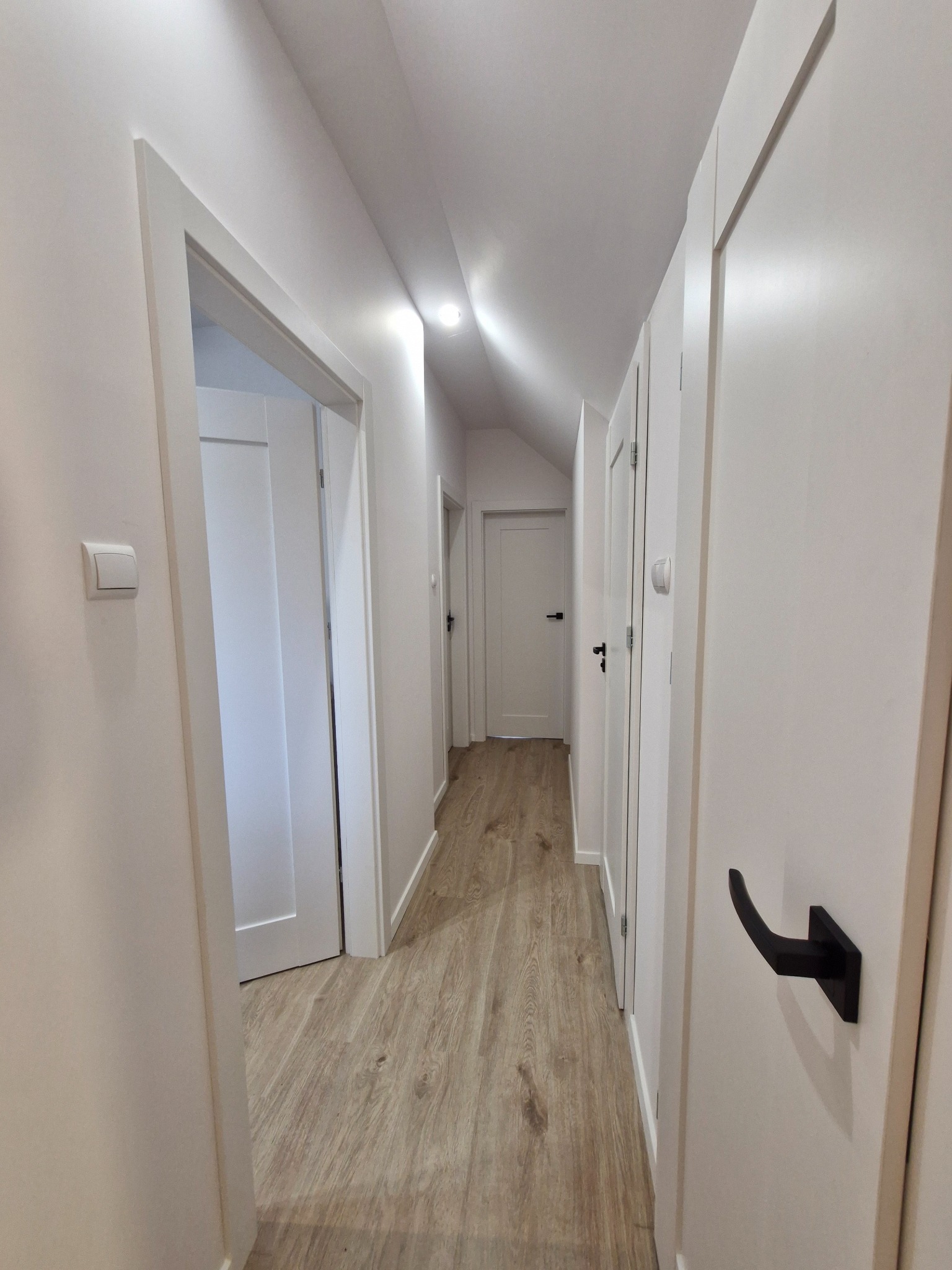 Korytarz po remoncie z jasnymi ścianami, panelami podłogowymi imitującymi drewno i białymi drzwiami z czarnymi klamkami.