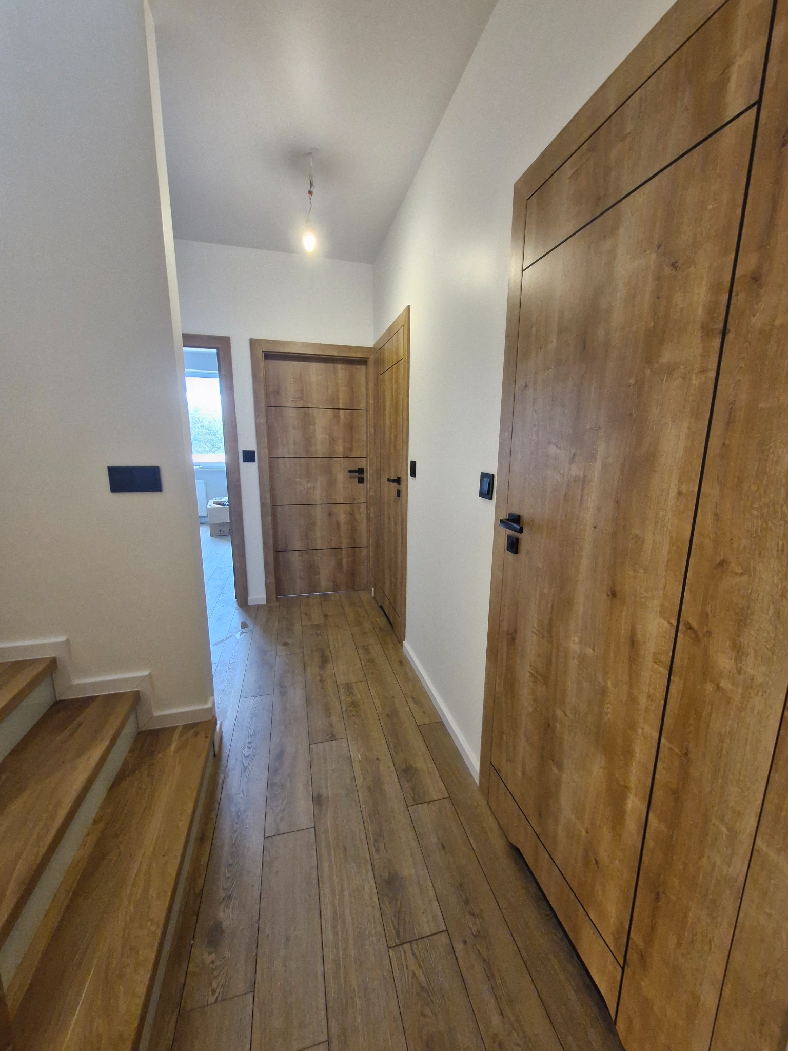 Korytarz po remoncie z drewnopodobną podłogą, schodami i dwiema parami drewnianych drzwi z czarnymi klamkami, oświetlony pojedynczą żarówką.
