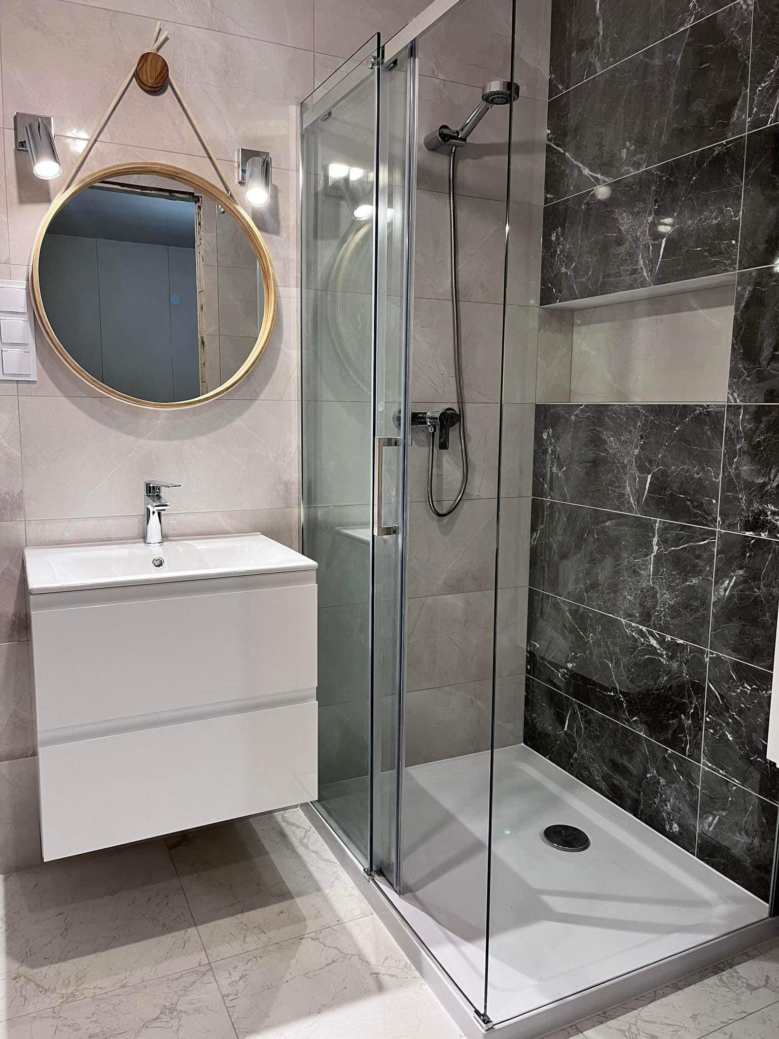 Nowoczesna łazienka z kabiną prysznicową, umywalką z szafką i okrągłym lustrem z drewnianym obramowaniem zawieszonym na sznurku, płytki w odcieniach szarości i czerni imitujące marmur.