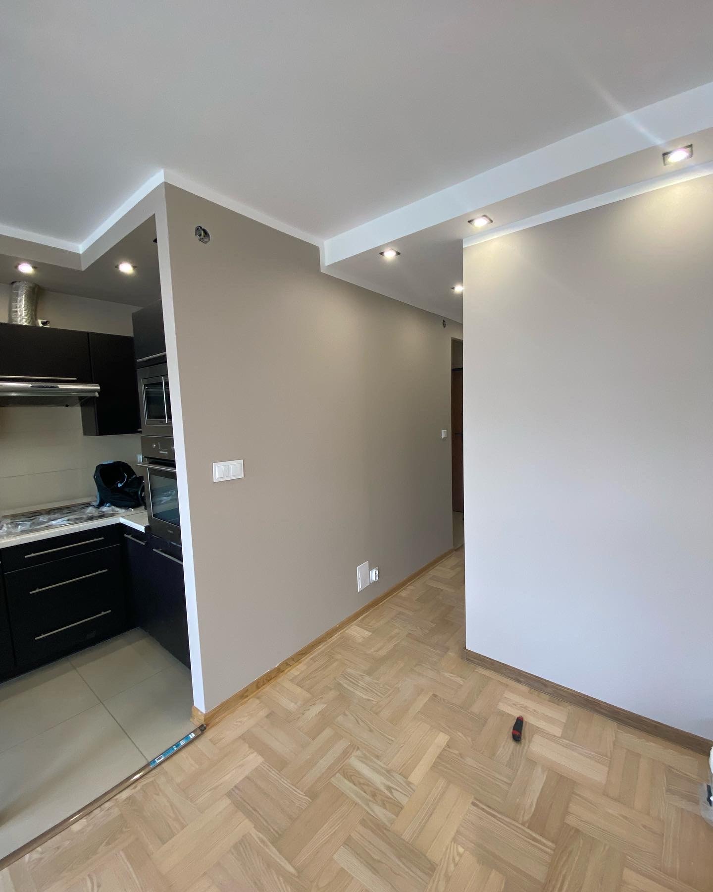 Wykończone wnętrze mieszkania z parkietem w jodełkę, świeżo pomalowanymi ścianami w odcieniach beżu i szarości oraz fragmentem zabudowy kuchennej z czarnymi szafkami i sprzętem AGD.