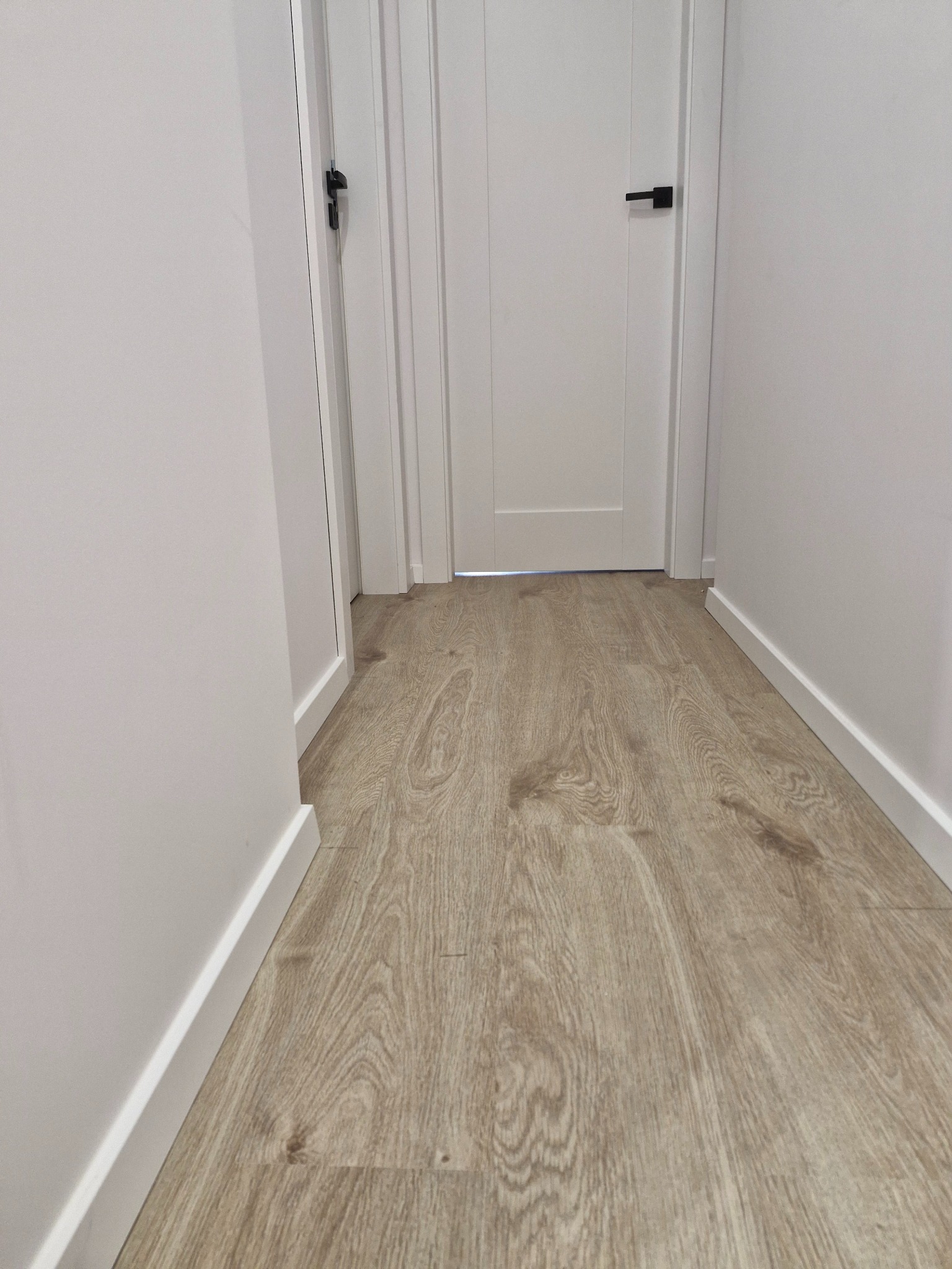Jasne panele podłogowe imitujące drewno w korytarzu z białymi ścianami i białymi drzwiami z czarnymi klamkami.