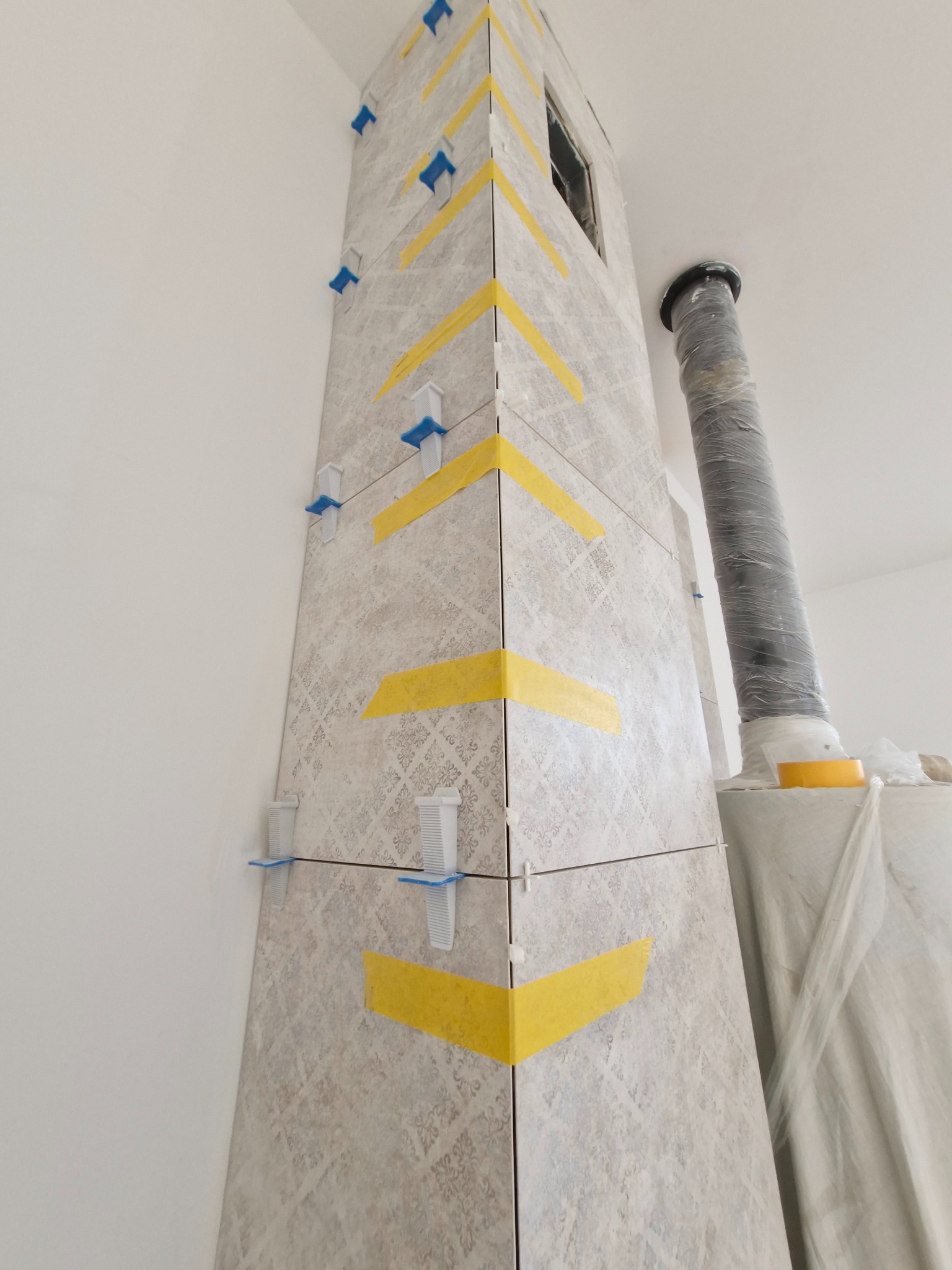 Wykończenie narożnika komina płytkami ceramicznymi z użyciem poziomujących klipsów i taśmy malarskiej, widoczny obok rura wentylacyjna zabezpieczona folią.