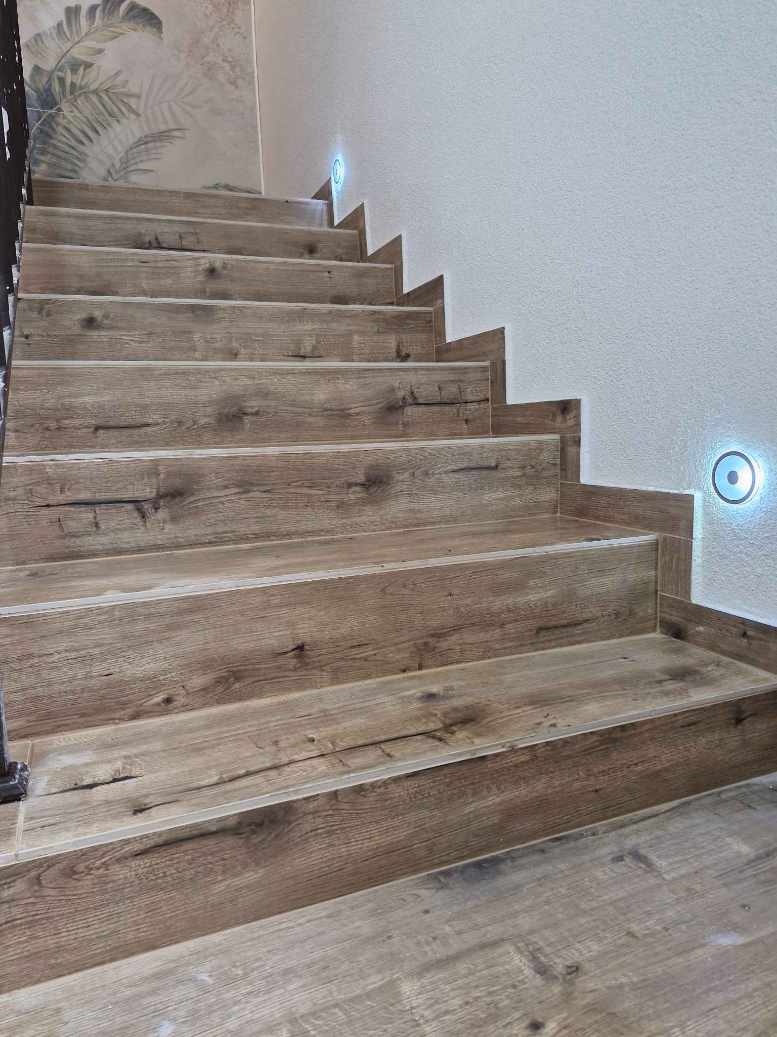 Schody wyłożone płytkami imitującymi drewno, z oświetleniem punktowym na ścianie i dekoracyjną tapetą z motywem roślinnym w górnej części ściany.