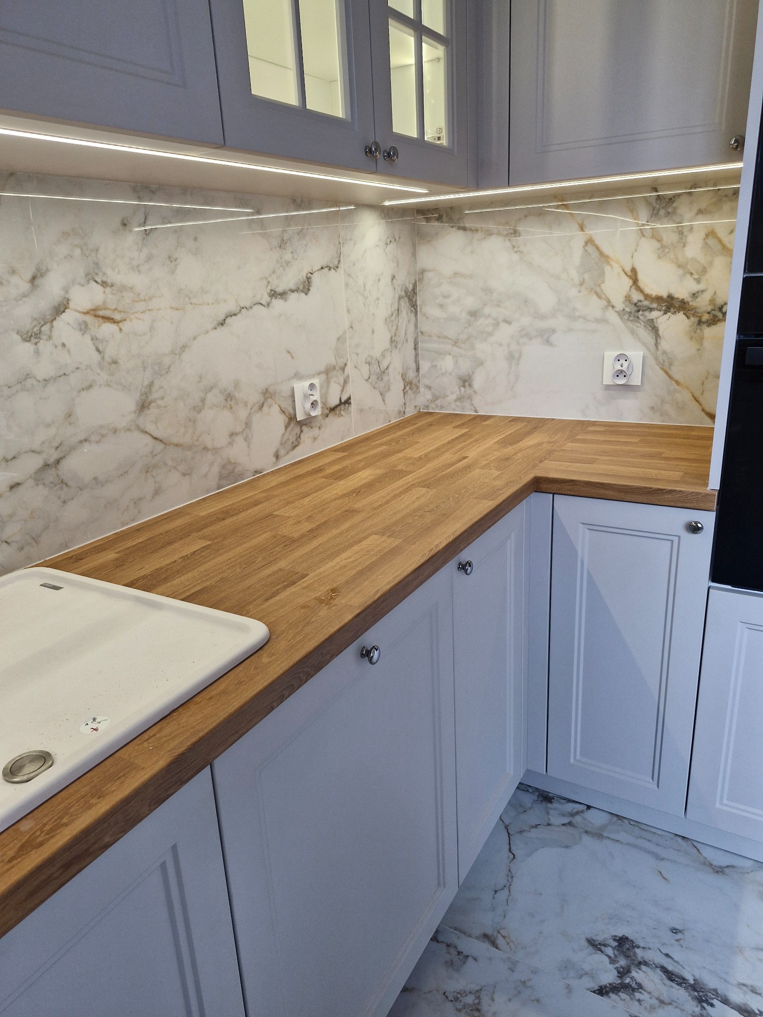 Jasna kuchnia z drewnianym blatem, marmurową ścianą i podświetleniem LED pod szafkami górnymi, widoczny zlew ceramiczny i gniazdka elektryczne.